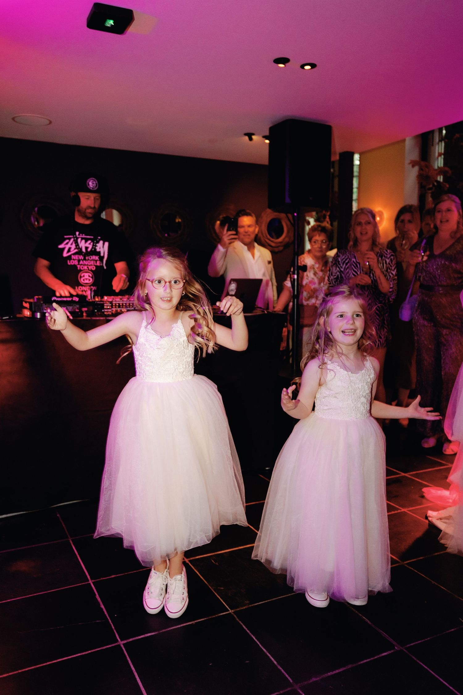 Two young girls in white tulle dresses dance together on a dark dance floor under pink lighting at an event.