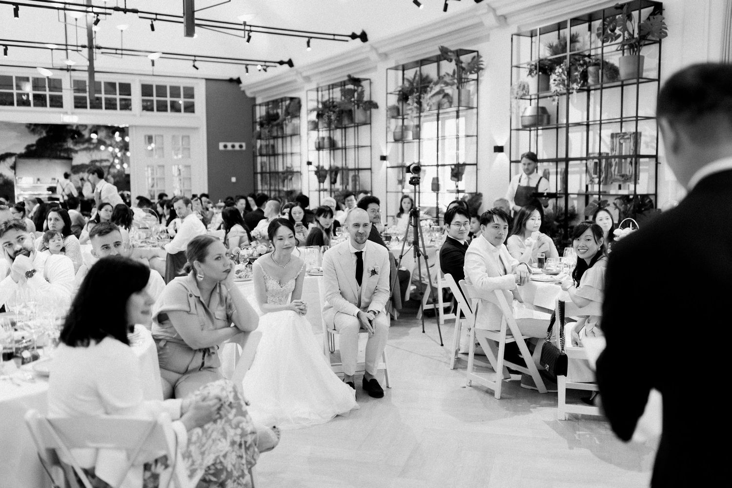Black and white photograph of a busy restaurant dining room with string lights and large windows during a dinner service.