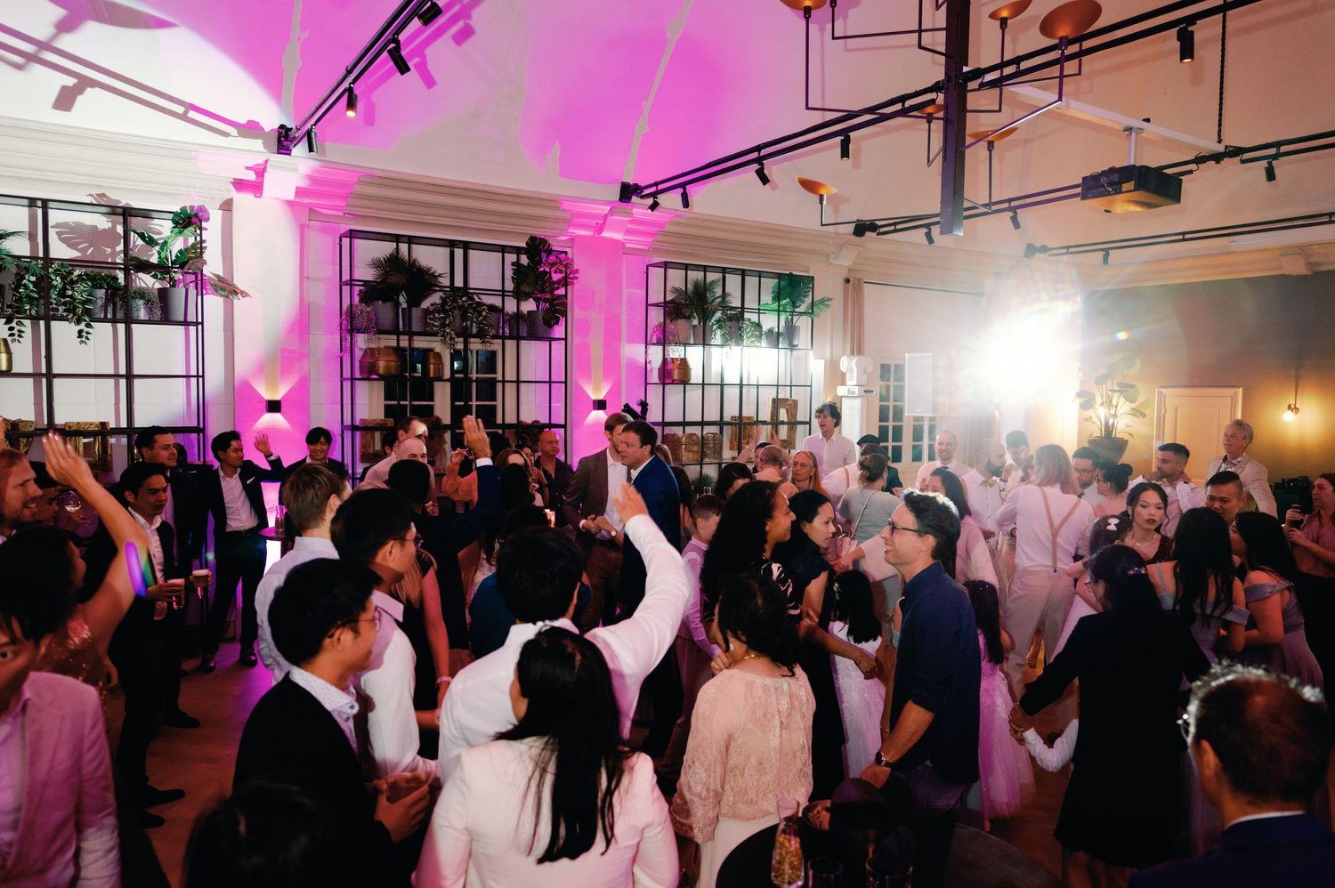 A crowded dance floor at a wedding reception with pink uplighting illuminating the modern industrial venue space.