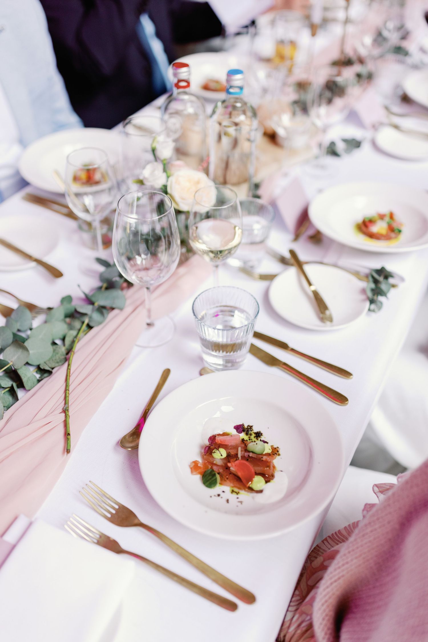 Elegant wedding reception table settings with white plates, gold cutlery, and eucalyptus greenery on a blush pink tablecloth.