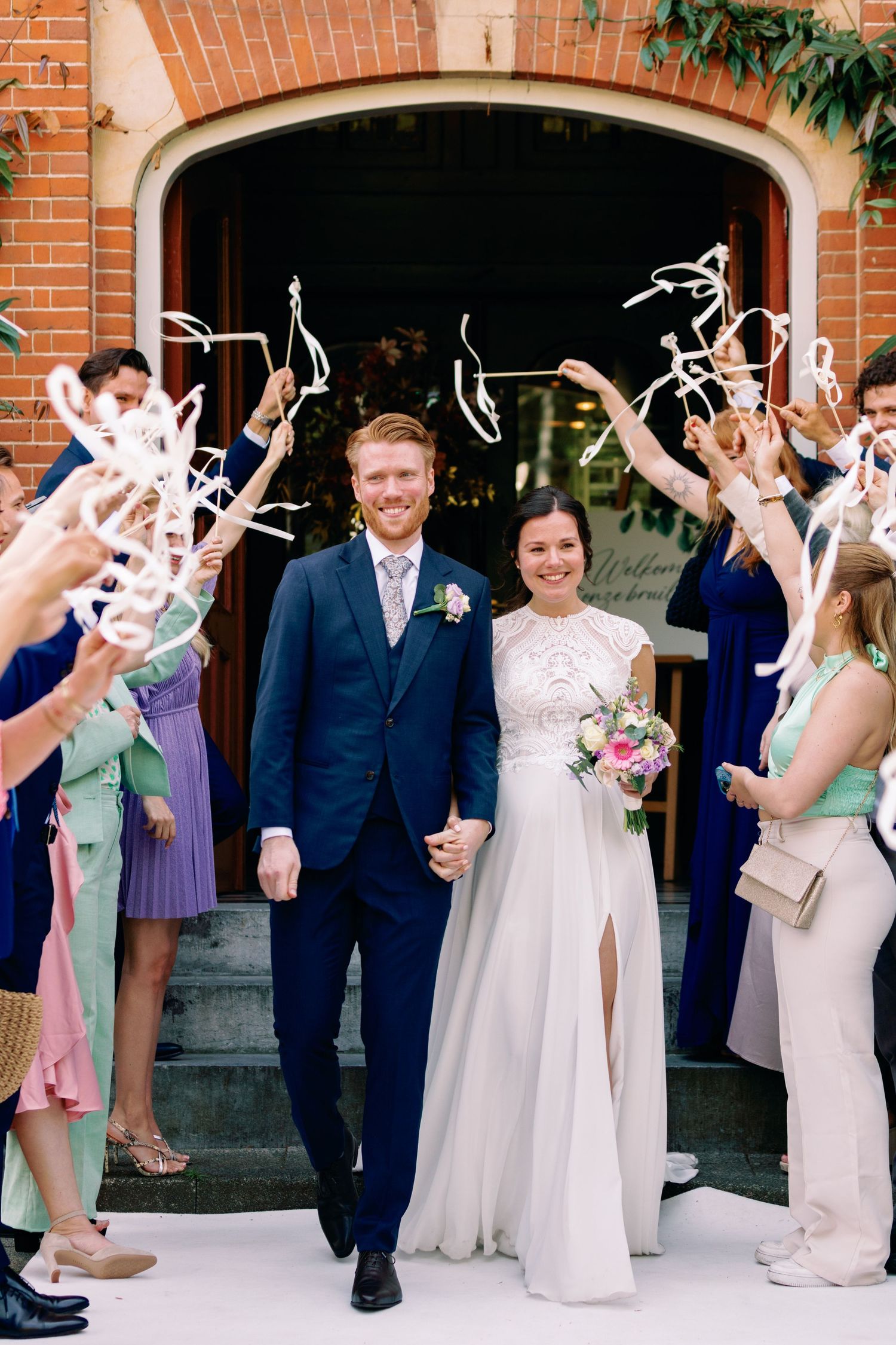 Wedding party in formal attire celebrates with white ribbon wands outside brick church entrance.