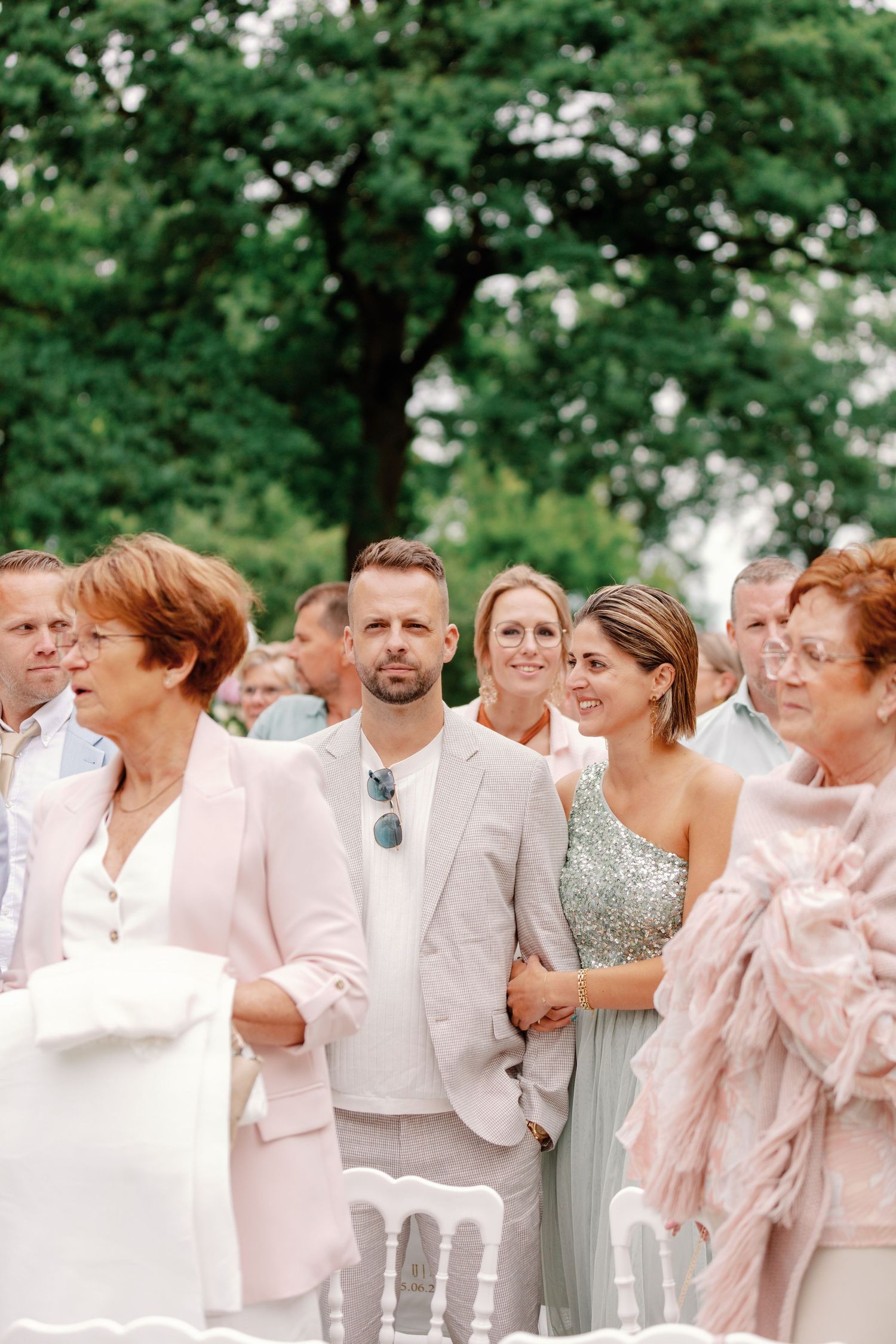 Wedding party guests dressed in light pink and gray attire gather outdoors for group photos in a garden setting.