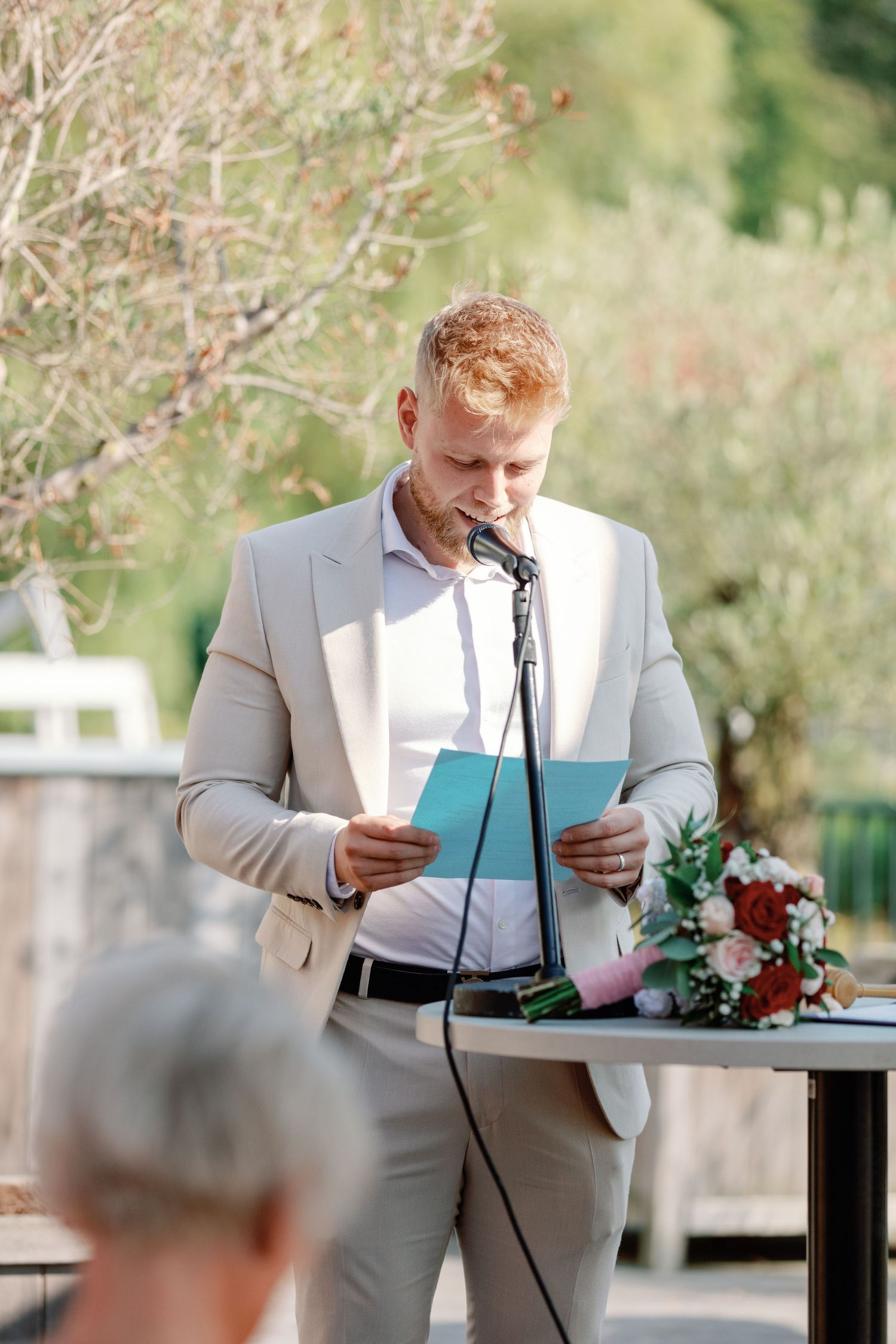 A speaker in a light beige suit reads from papers at an outdoor wedding ceremony.