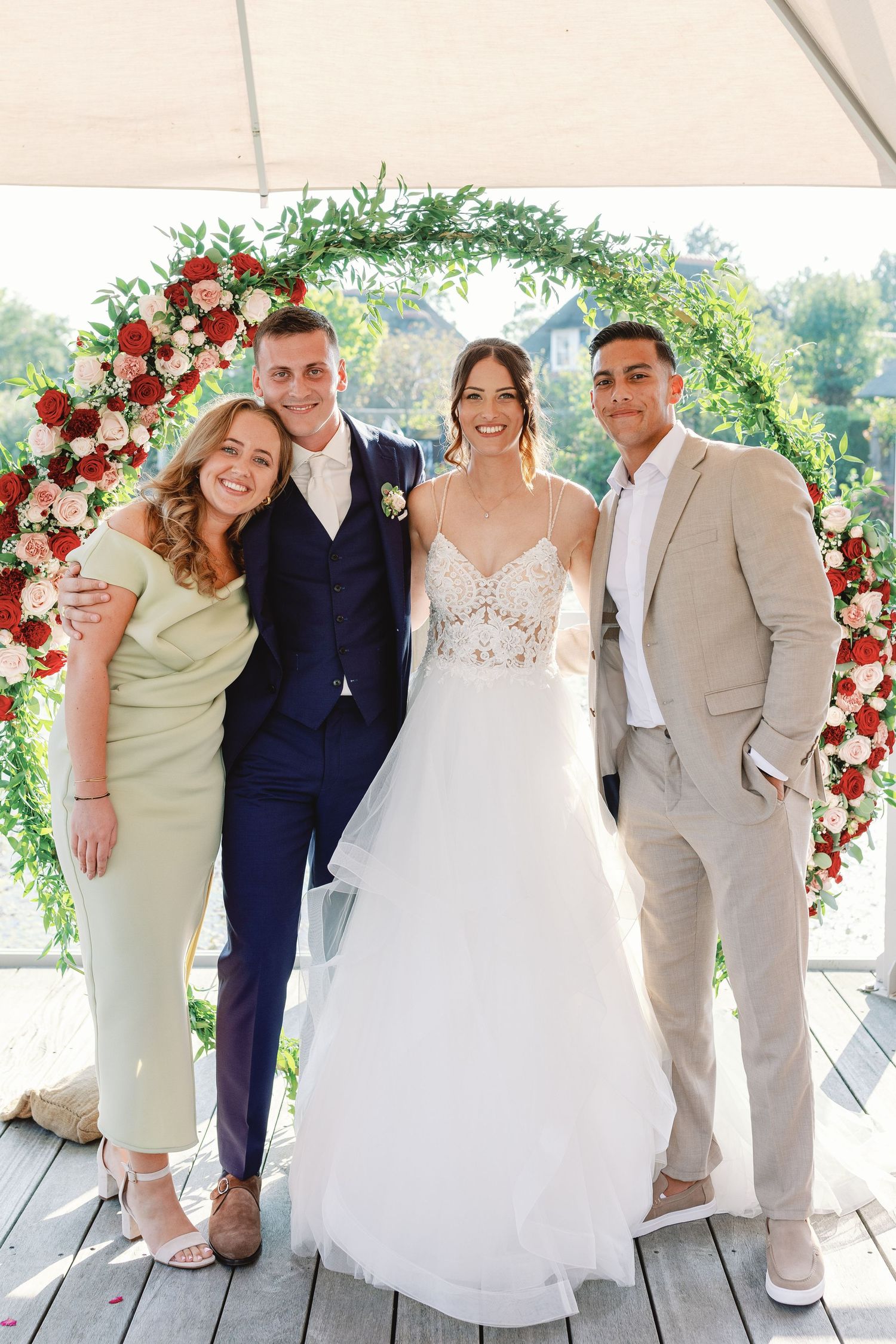 Wedding party poses together under decorated arch with red and white flowers.