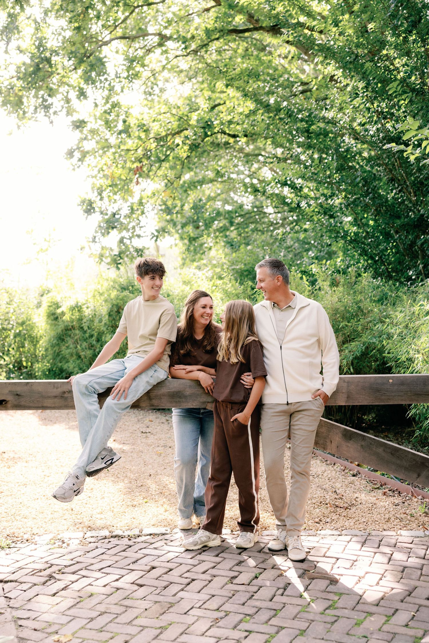Family of four posing outdoors on a brick path beside a wooden fence, with a teen boy seated and the others standing under green trees.