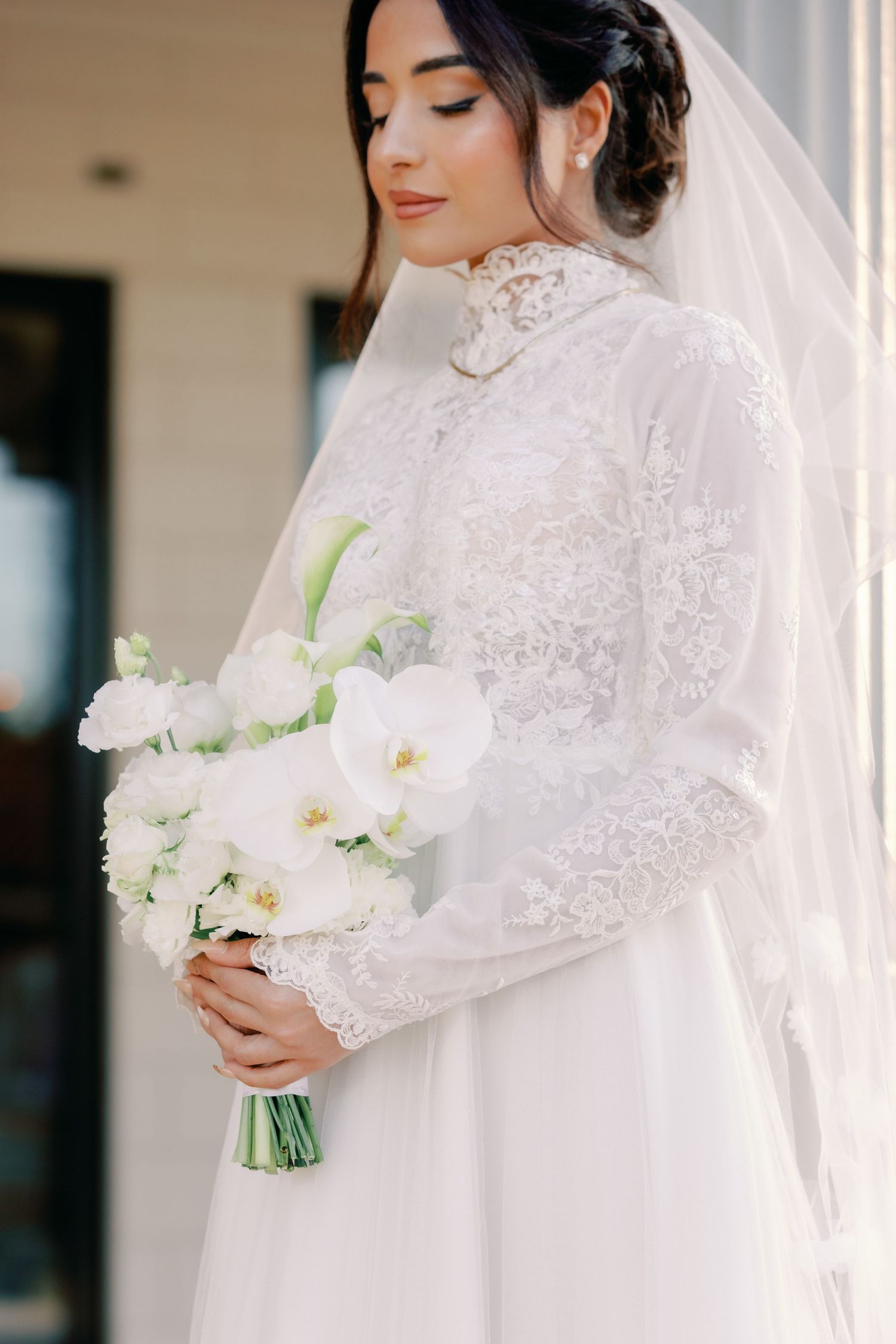 A bride in an elegant white lace gown with high neck and long sleeves holds a bouquet of white flowers on her wedding day.