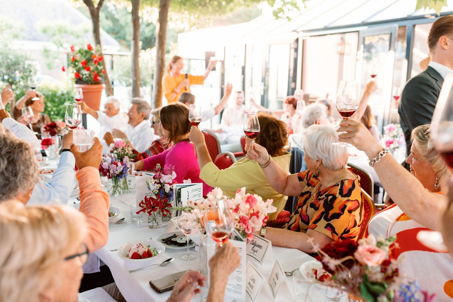 Guests raising wine glasses in a toast at an elegant outdoor wedding reception with floral centerpieces.