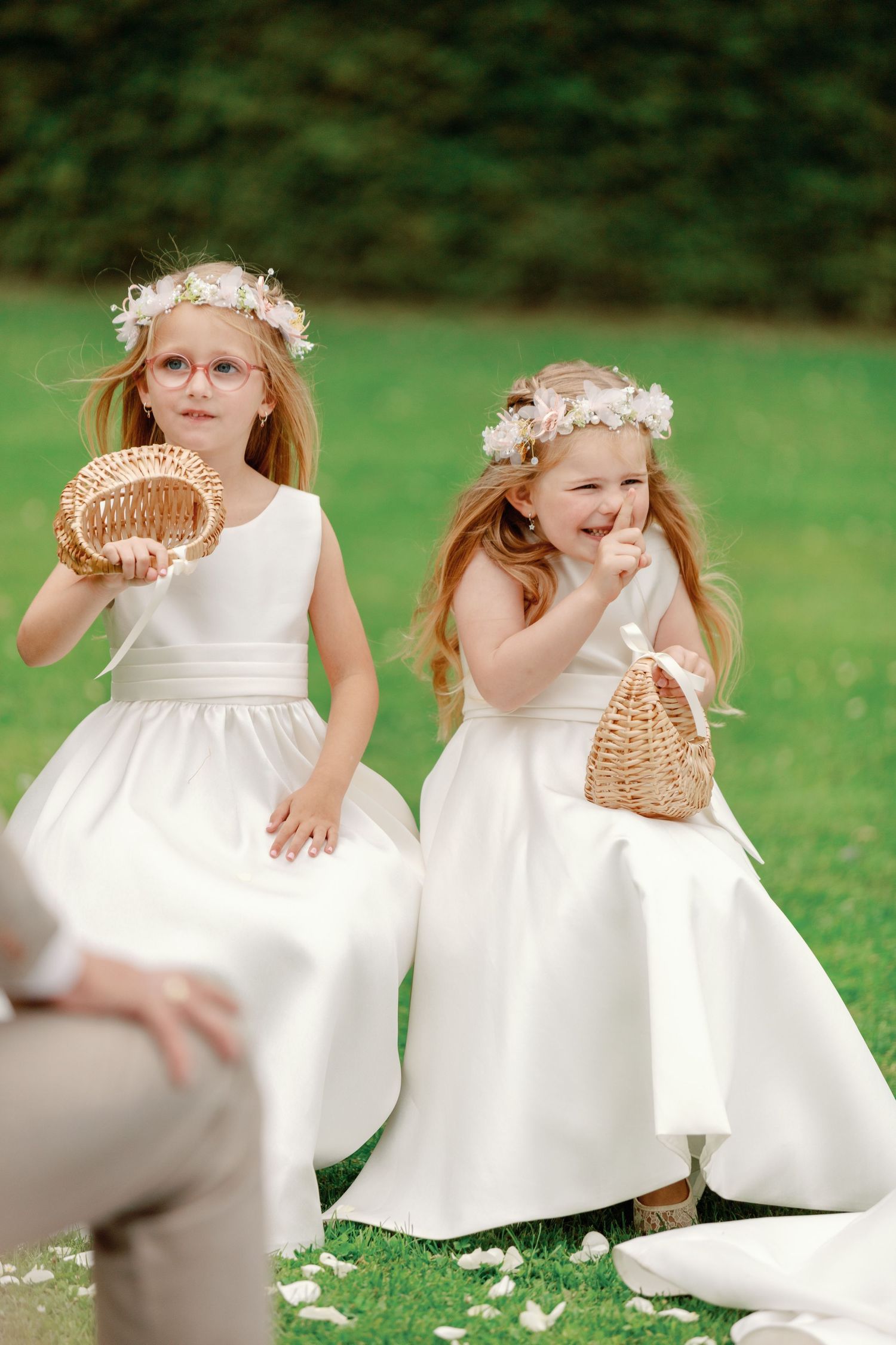 Two flower girls in white dresses and floral crowns carry woven baskets on a grassy lawn at a wedding.