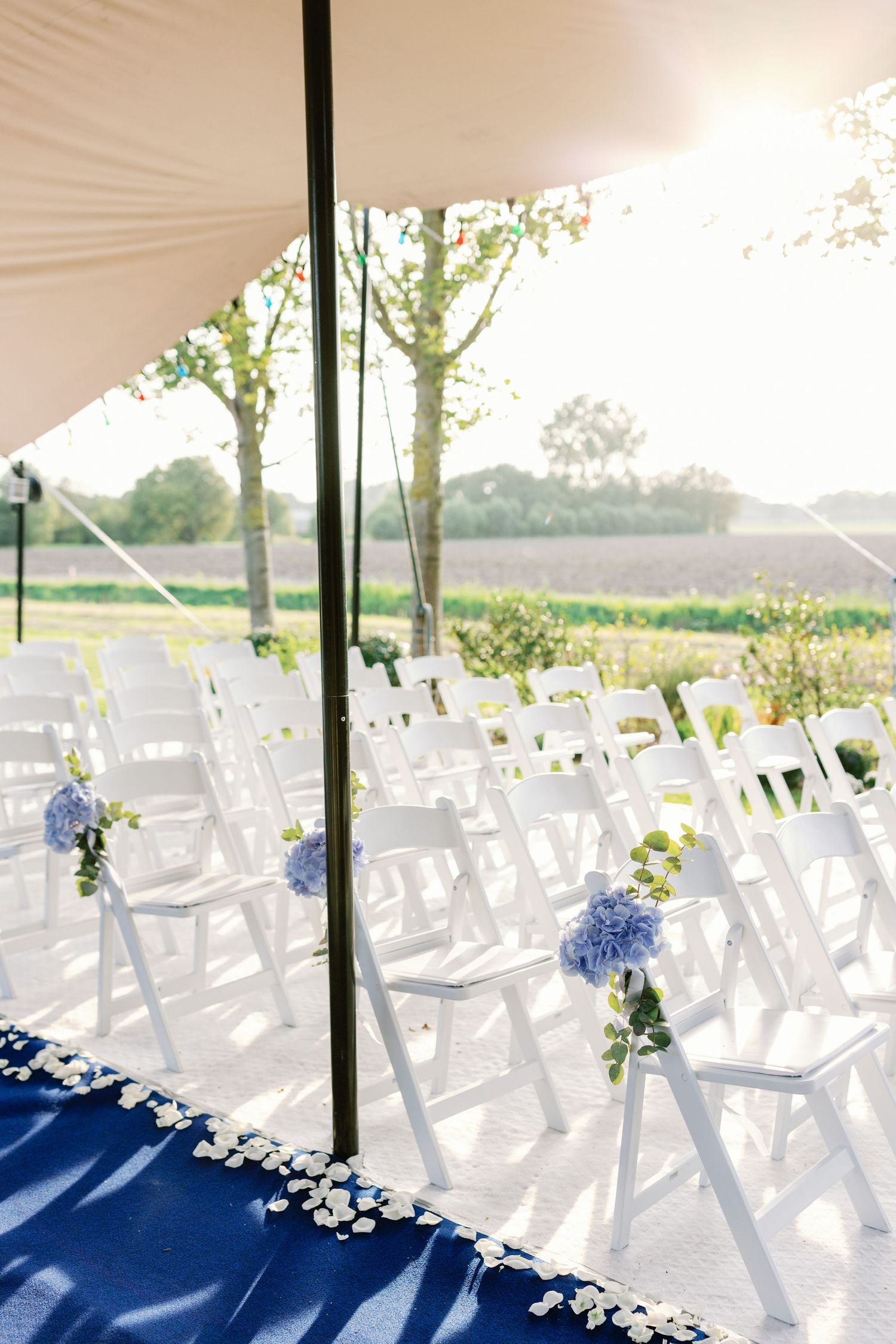 White ceremony chairs set up under draped tent with blue floral aisle decorations.