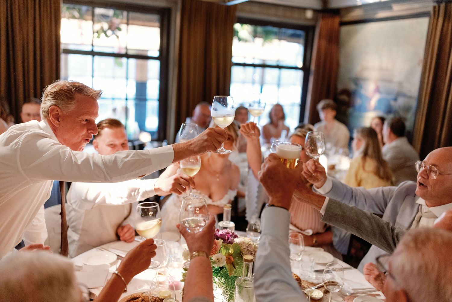 Wedding guests raise their glasses in a celebratory toast during a reception at a rustic venue.
