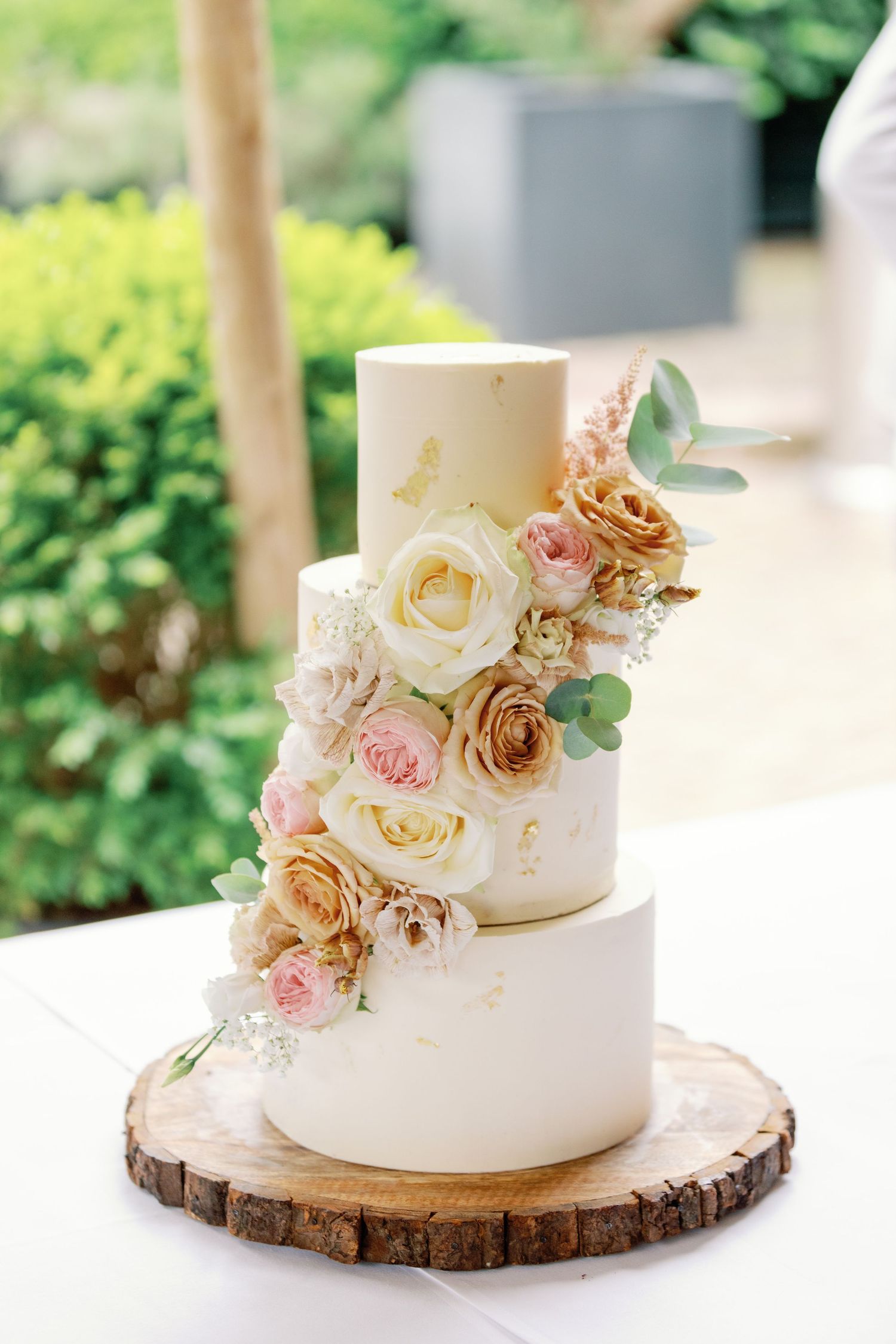 Elegant two-tier white wedding cake adorned with blush and caramel roses on rustic wood slice cake stand.