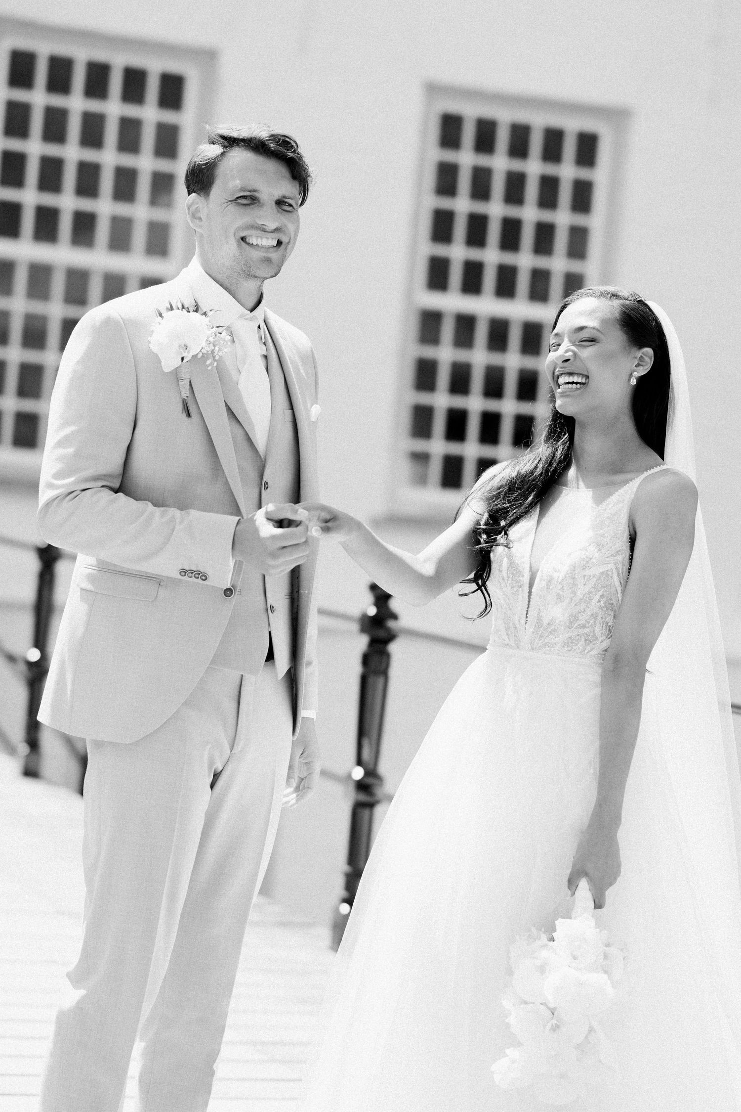 A happy couple laughs together in formal wedding attire during an outdoor photoshoot in black and white.