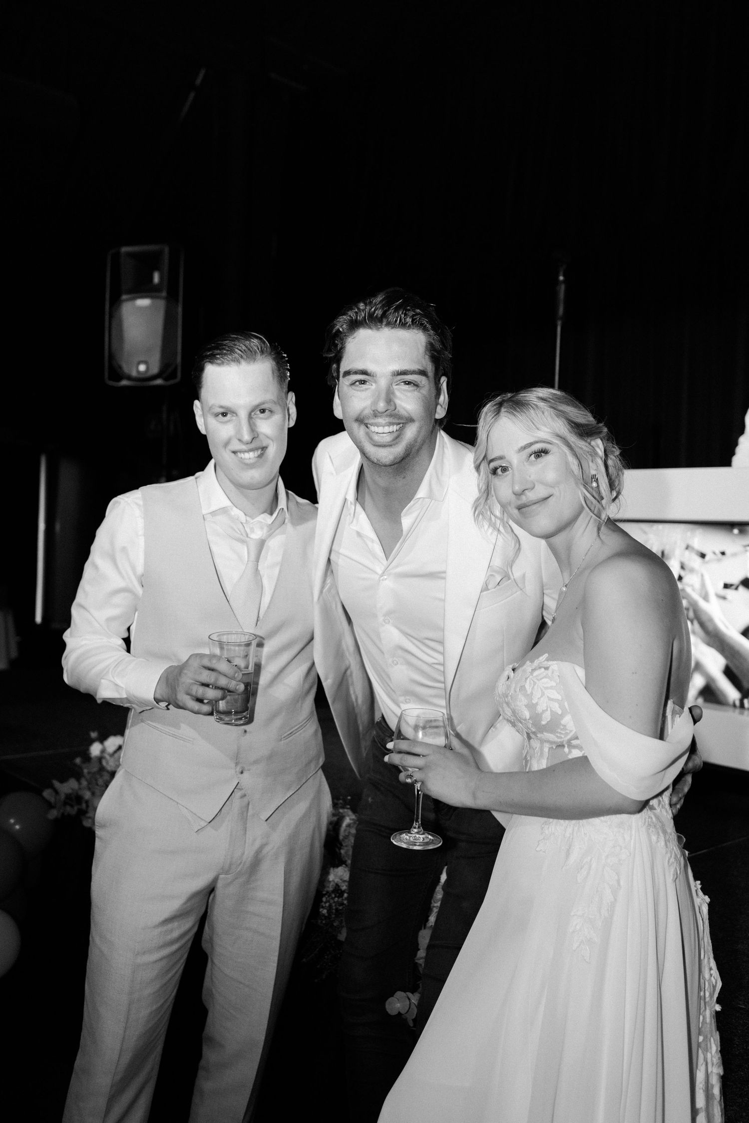 Wedding party members in light colored attire share laughter and drinks during an evening reception celebration.