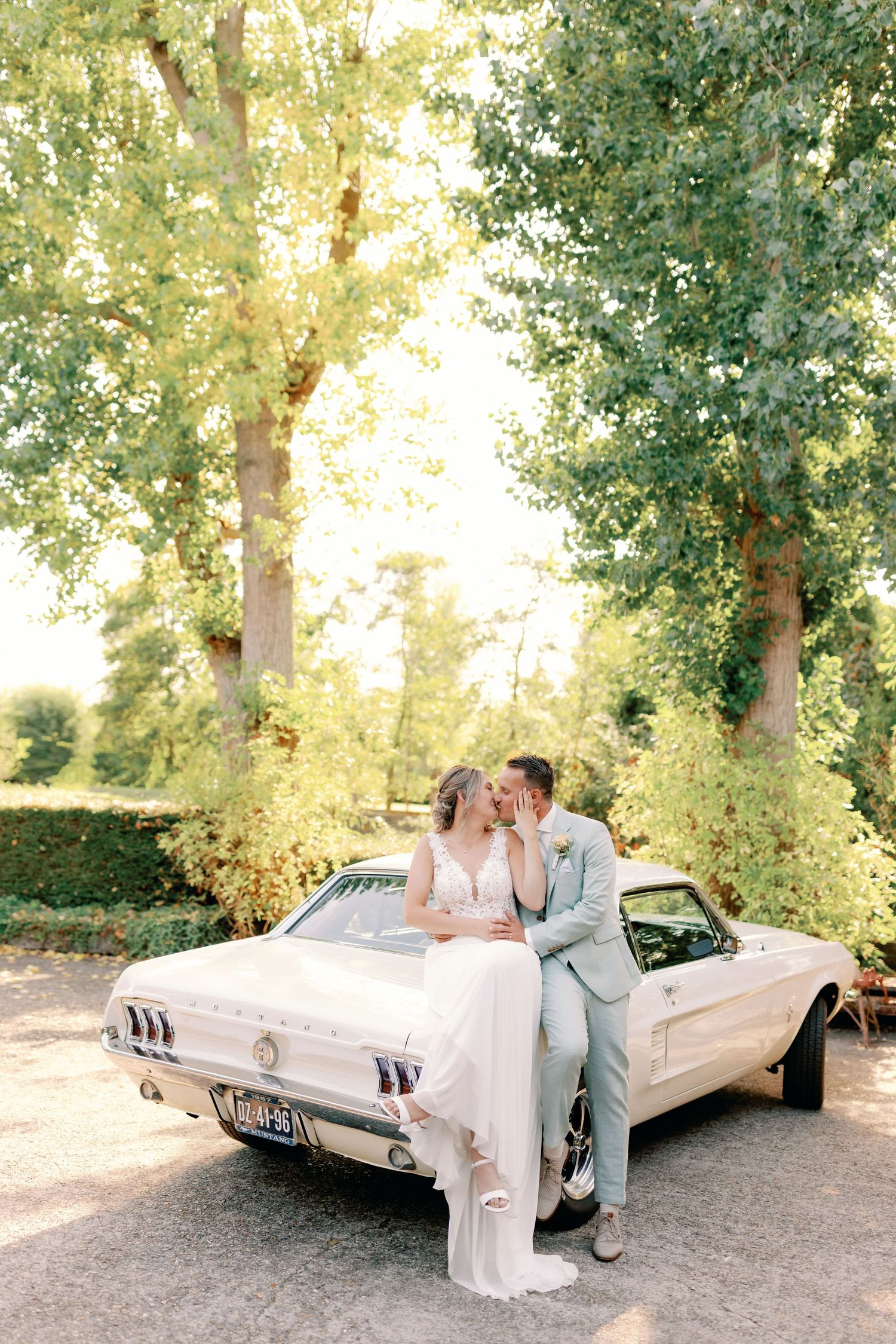 A vintage white car parked in a sun-dappled grove provides a stylish setting for an intimate moment.