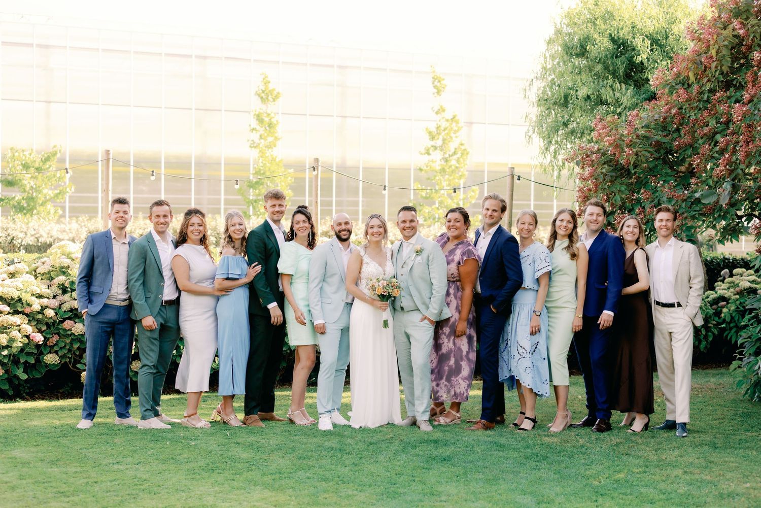 A group of wedding guests and newlyweds pose together on a sunny lawn with trees and greenery in the background.