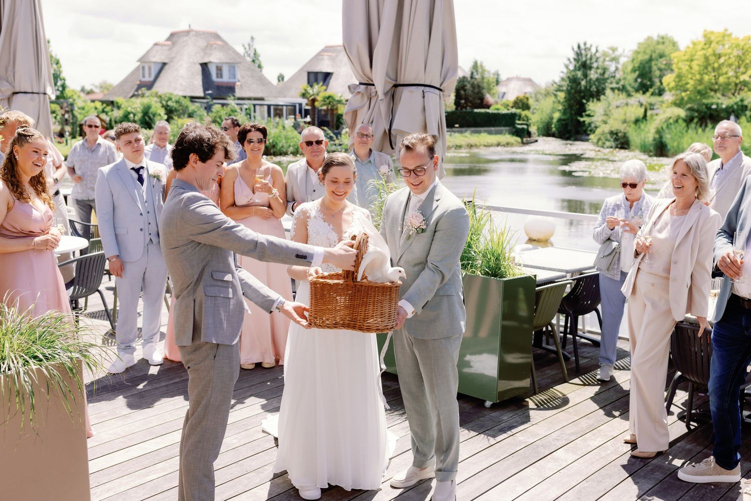 Bride and groom receive a wicker basket gift from a guest during an outdoor wedding reception by a scenic pond.