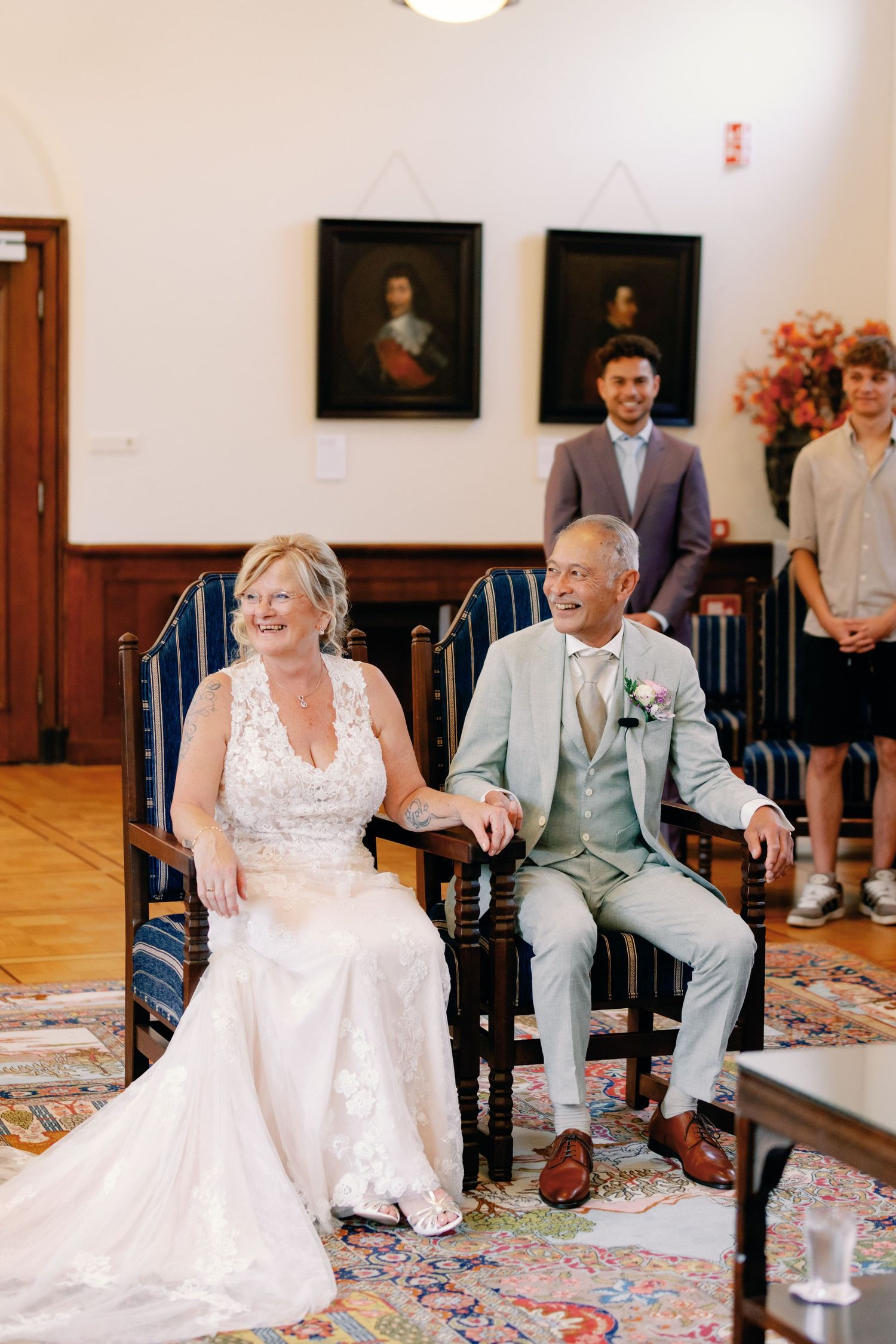 A joyful bride and groom sit together laughing during their wedding ceremony in an elegant room with portrait paintings.