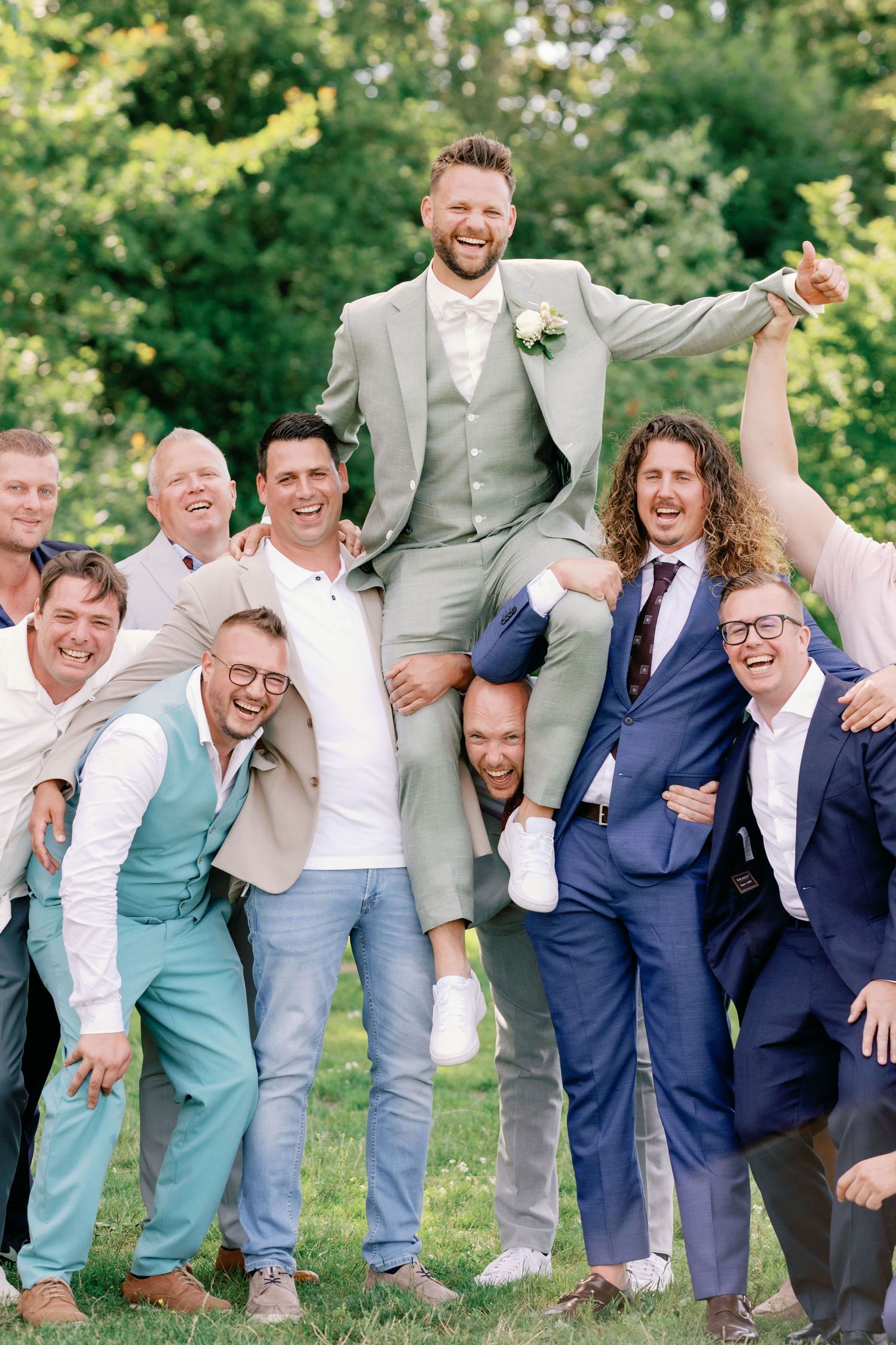 Groomsmen in navy and gray suits pose playfully in a line outdoors with trees in the background.