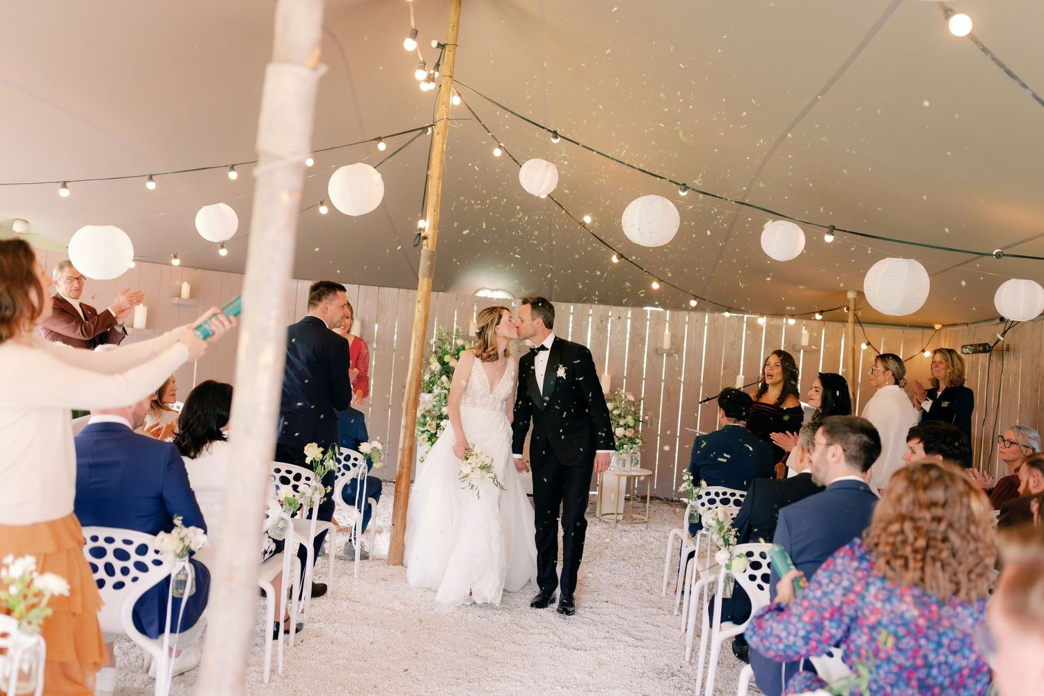 Bride and groom share a kiss at elegant tent wedding ceremony with white lanterns and confetti.