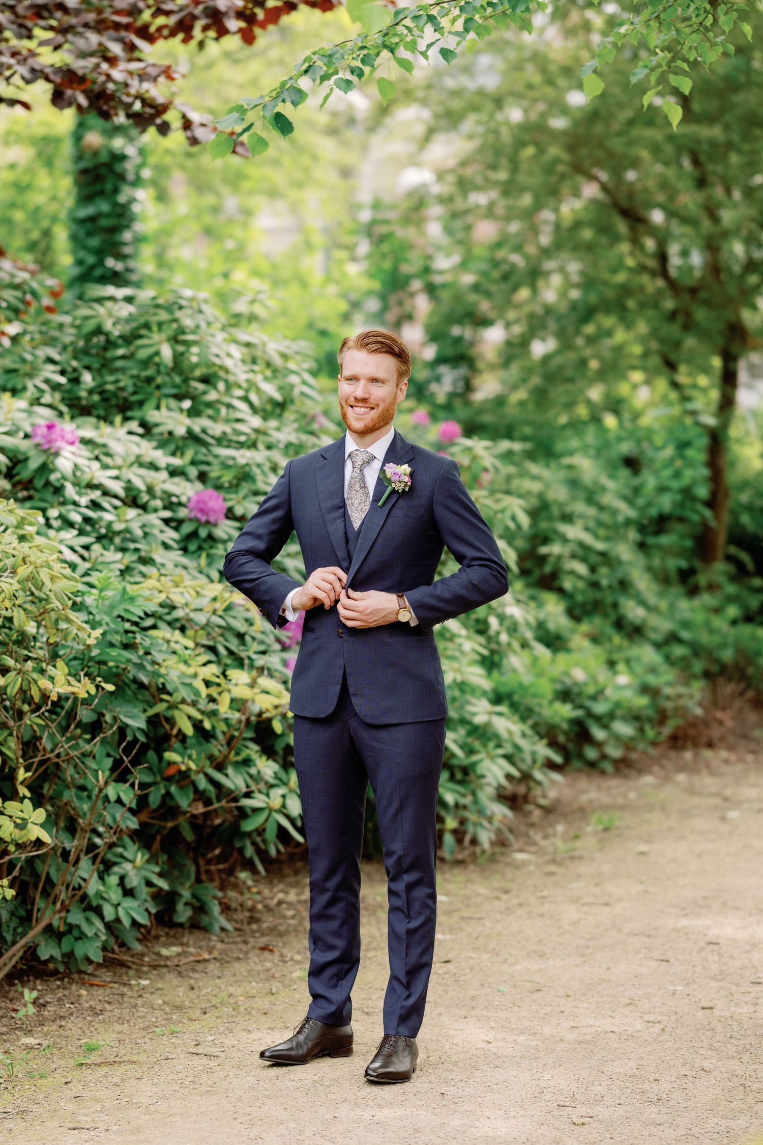 A person in a navy suit poses casually in a garden setting with lush greenery and flowers in the background.