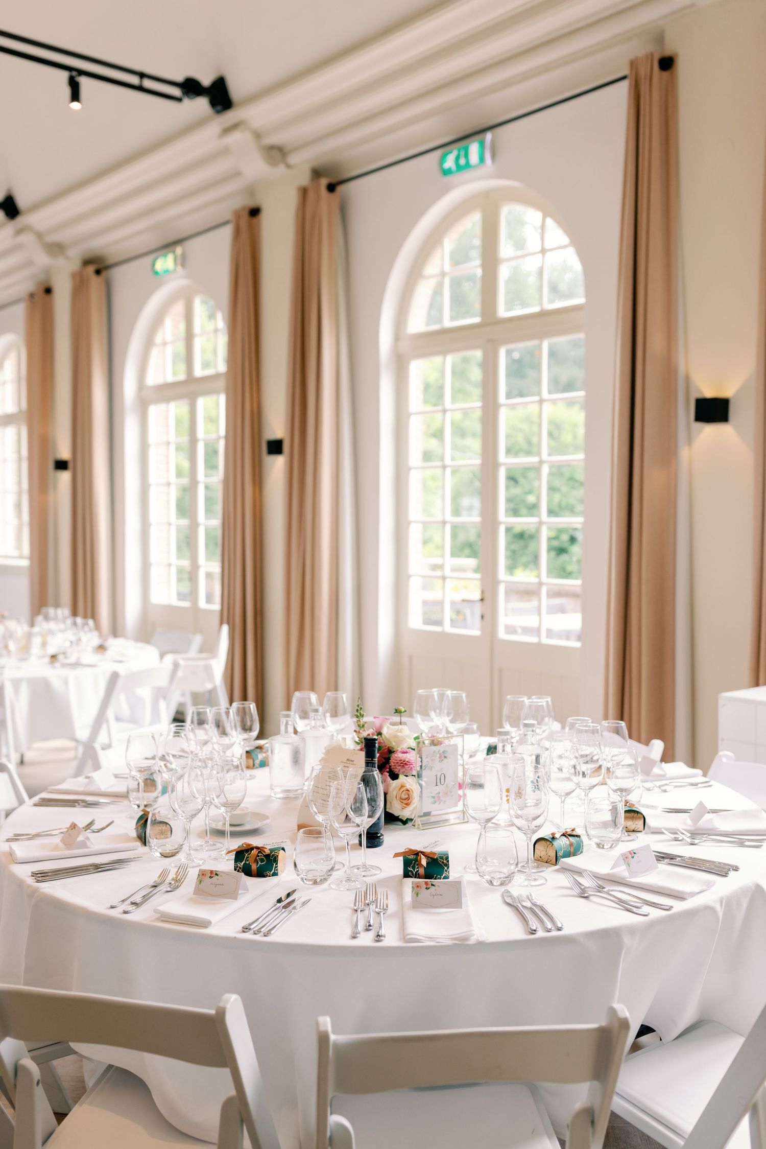 Elegant wedding reception table setting with white linens and glassware in a bright room with arched windows and beige curtains.