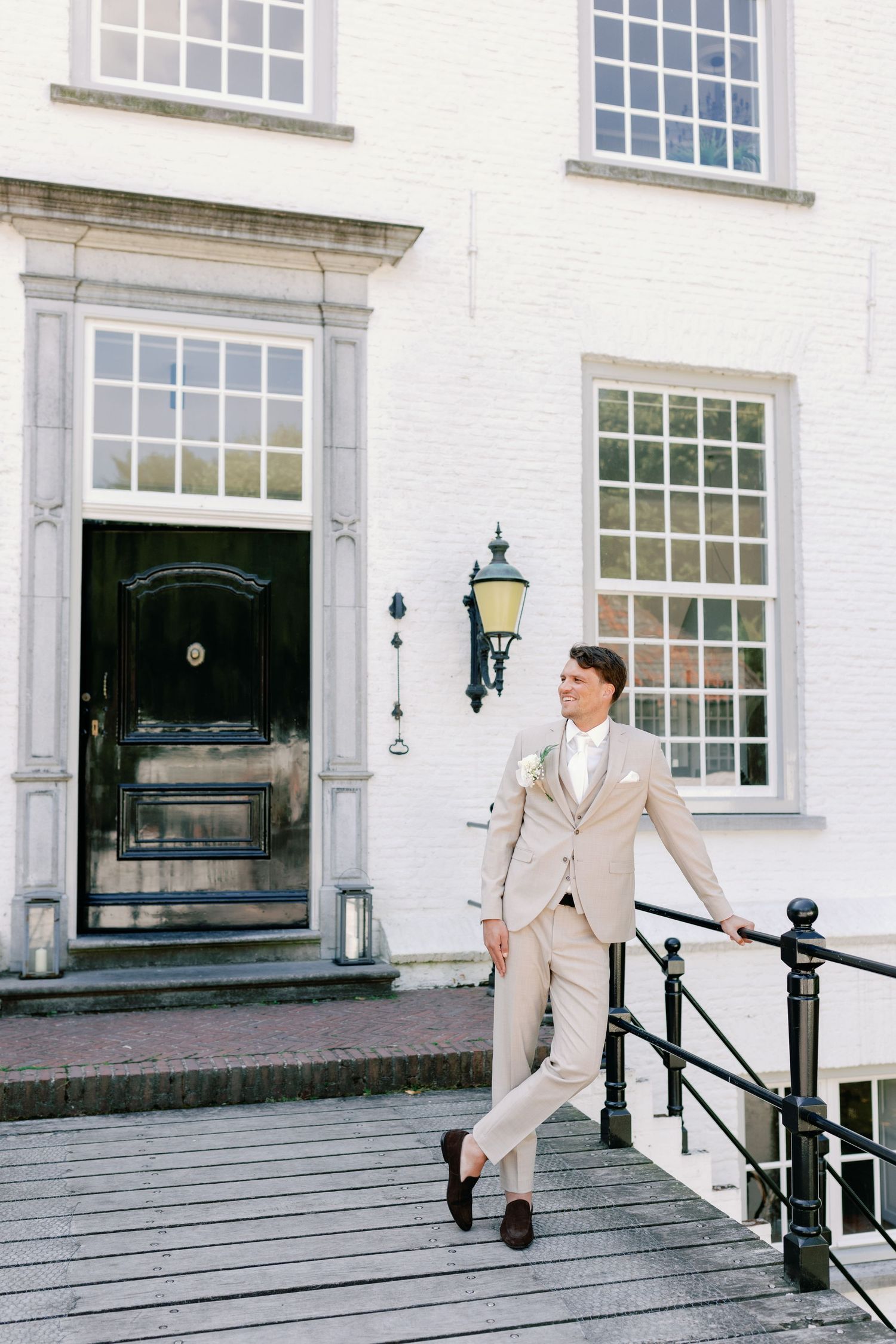 A person in a beige suit leans casually against black railings outside a white building with large windows.
