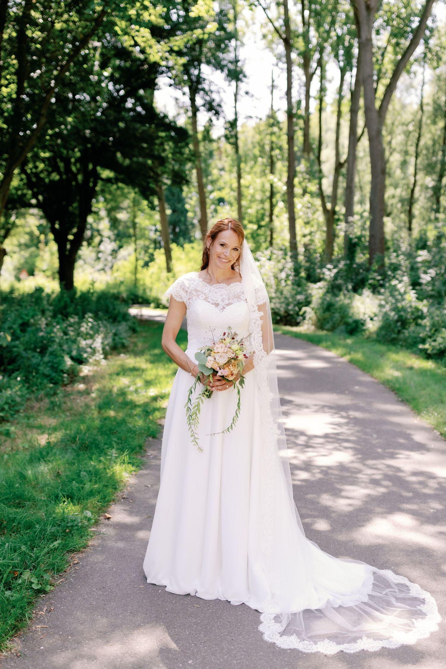 A bride in a white lace wedding dress poses on a tree-lined path holding a bouquet in a natural outdoor setting.