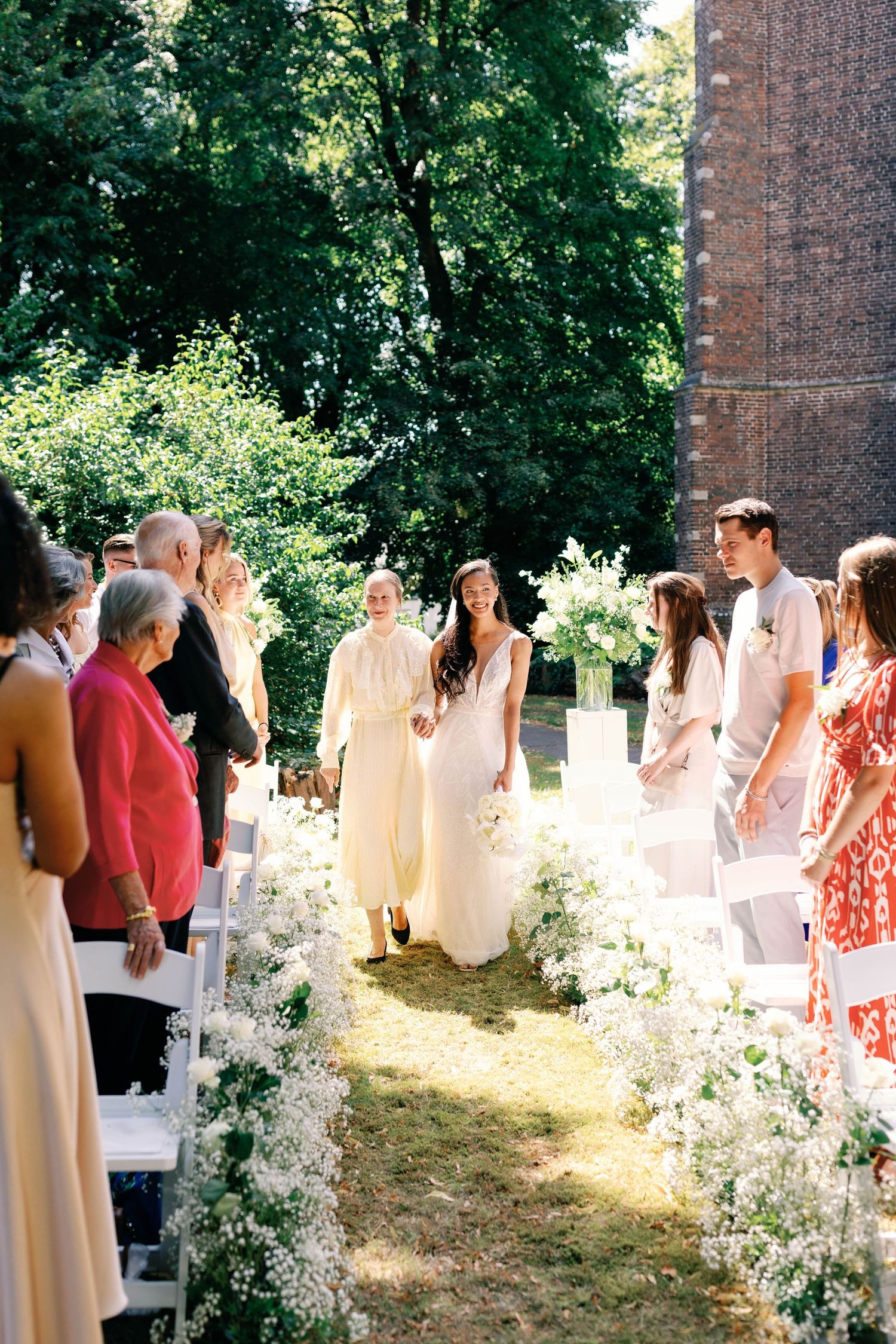 Wedding guests seated in white chairs outdoors with sunlit trees and brick building in background.