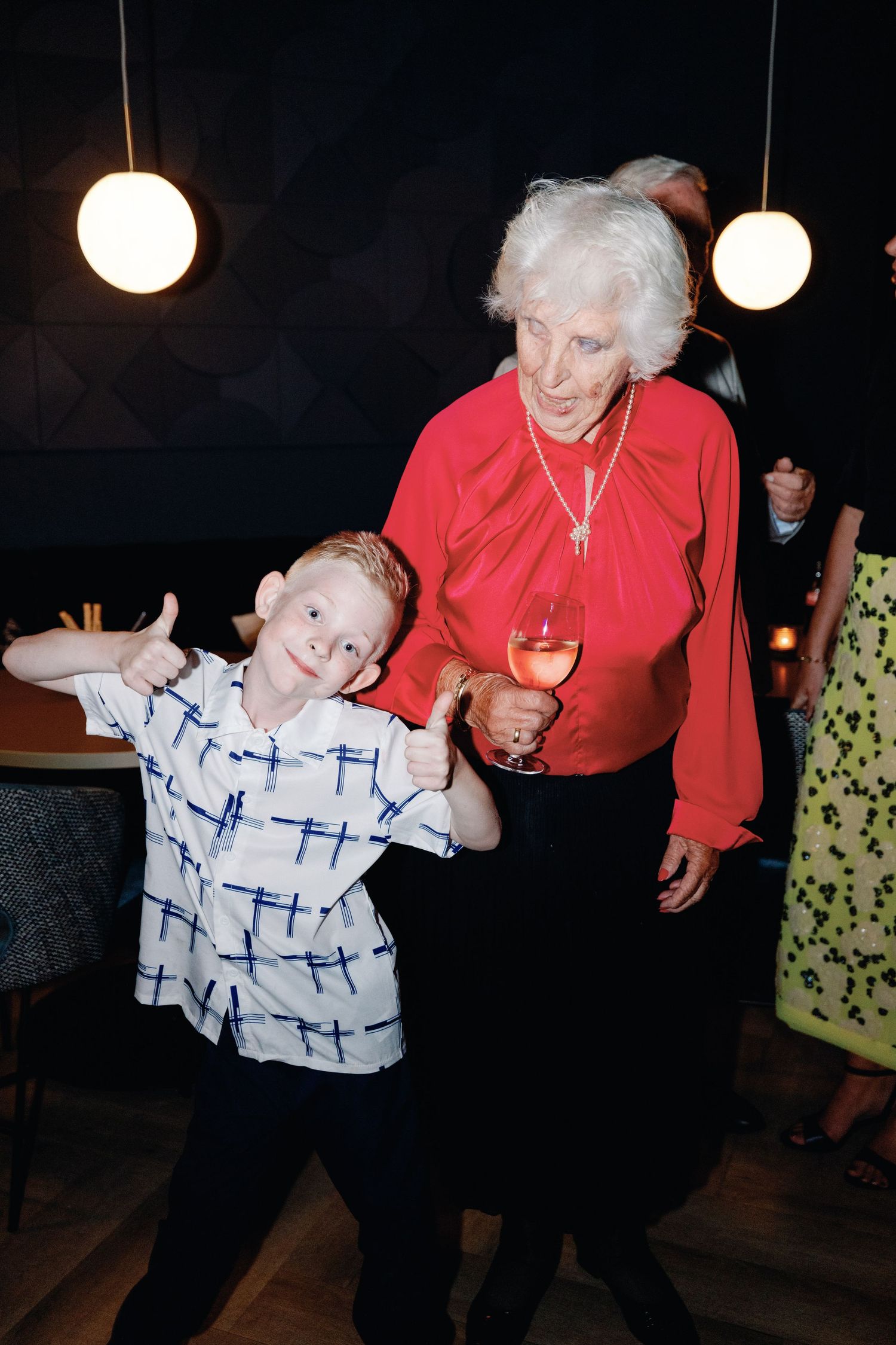 A young boy in a checkered shirt strikes a pose next to an elderly woman in red at a wedding reception.