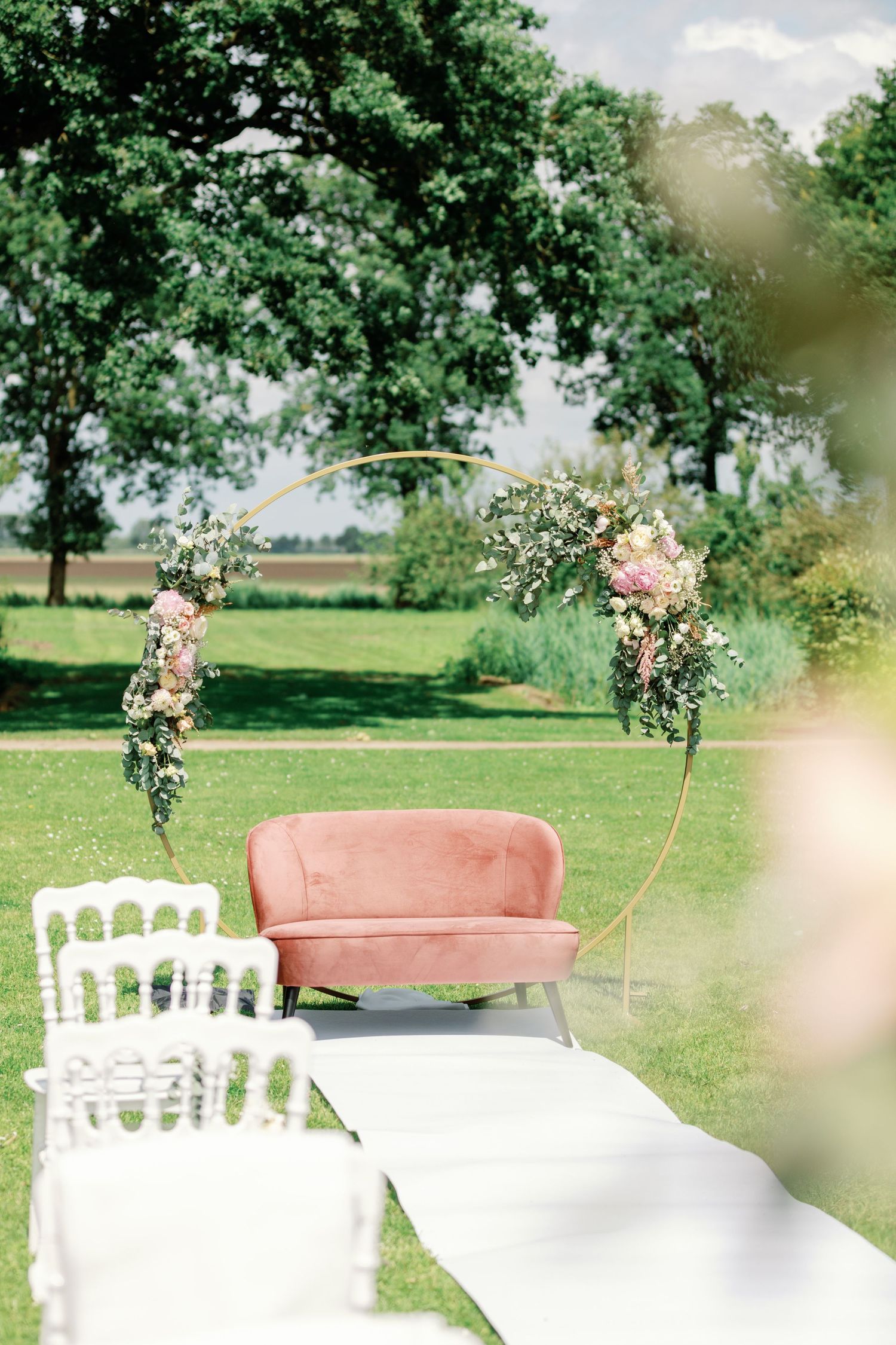 A pink vintage sofa and white chairs arranged outdoors on grass with floral decorations for a garden wedding ceremony.