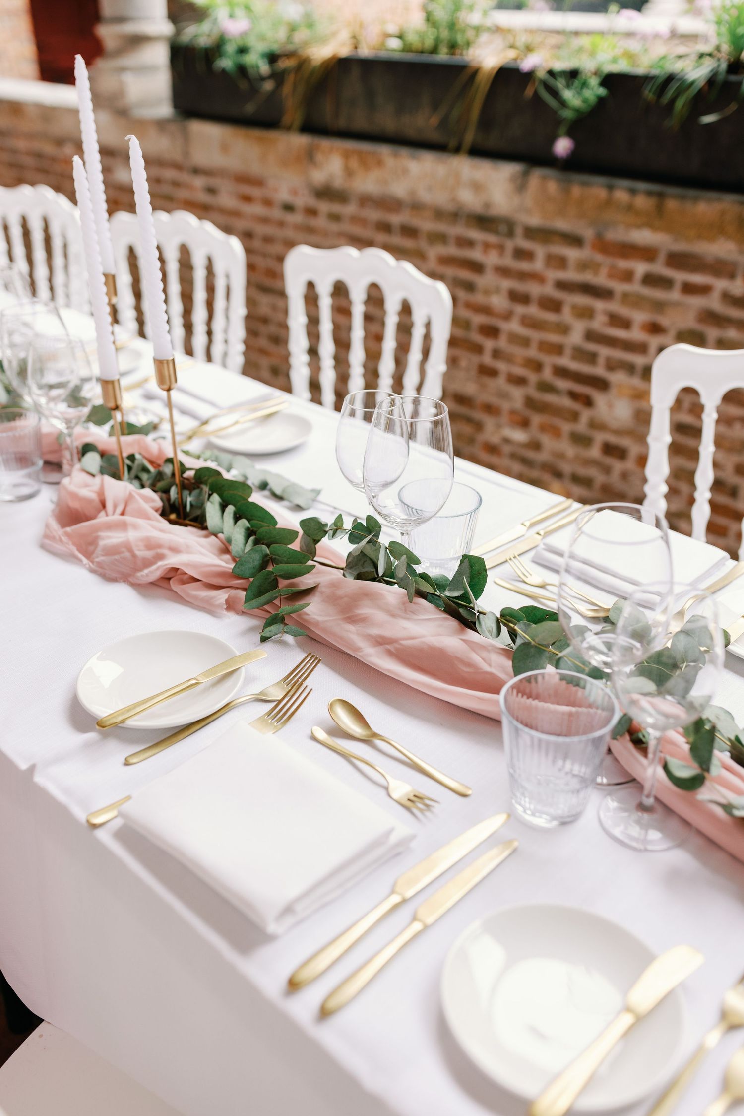 Elegant wedding table setting with white linens, gold cutlery, pink napkins and eucalyptus garland against exposed brick wall.