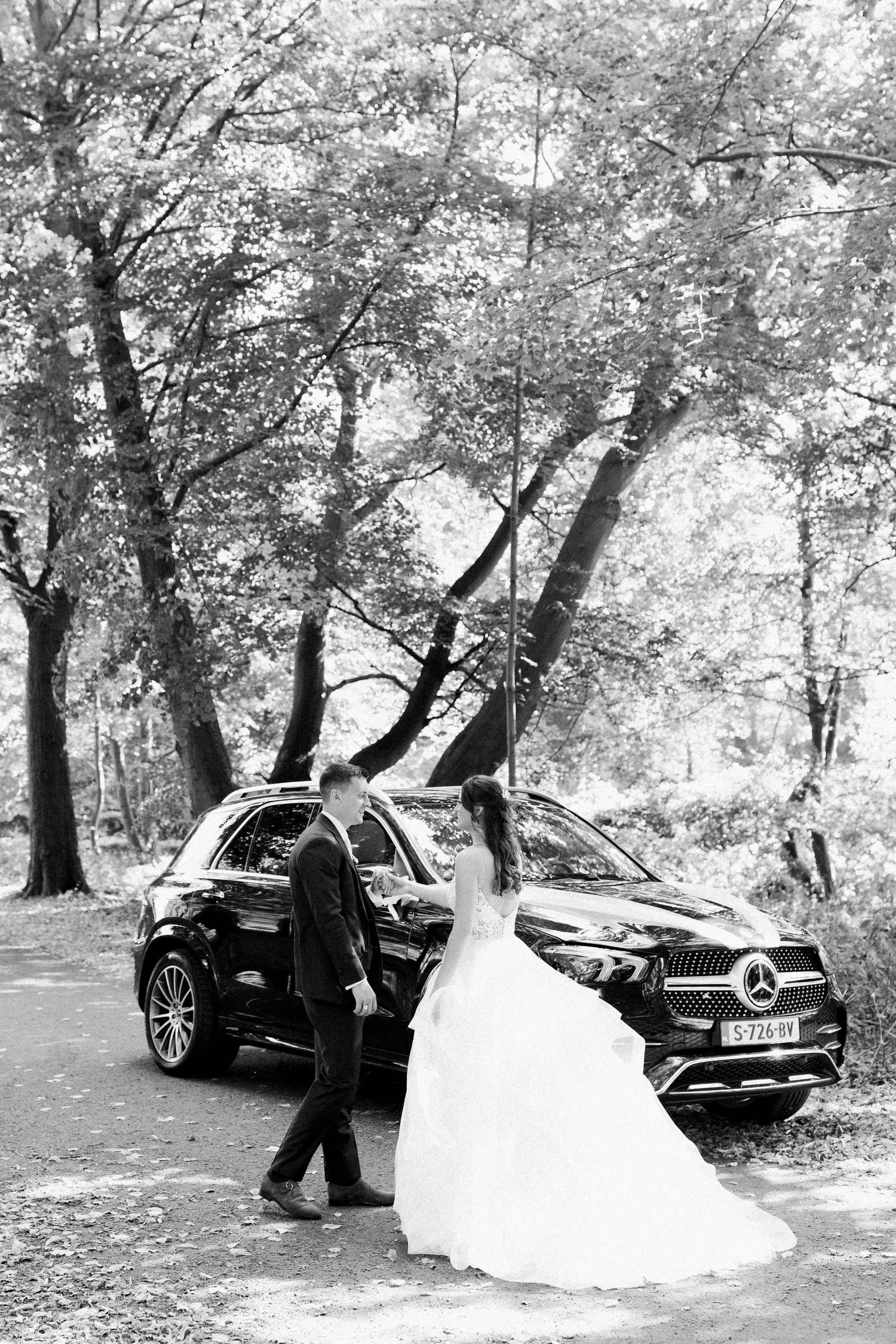 A black and white wedding photo featuring a Mercedes car in a wooded setting.