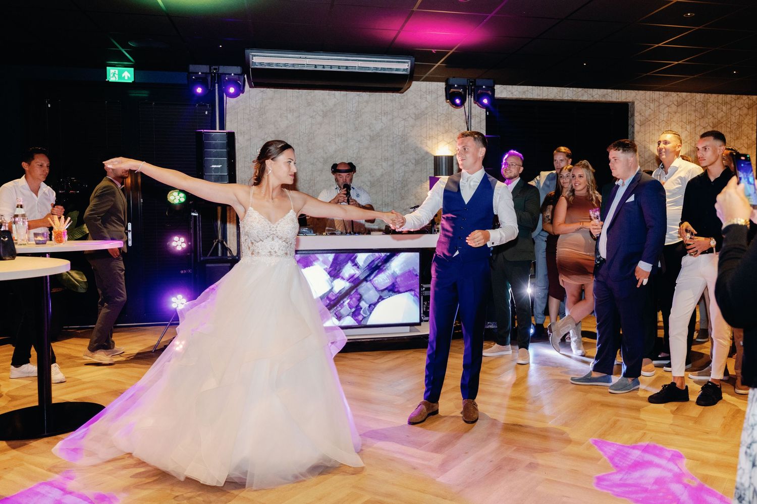 A couple dances energetically on a wooden dance floor under colorful party lights at their wedding reception.