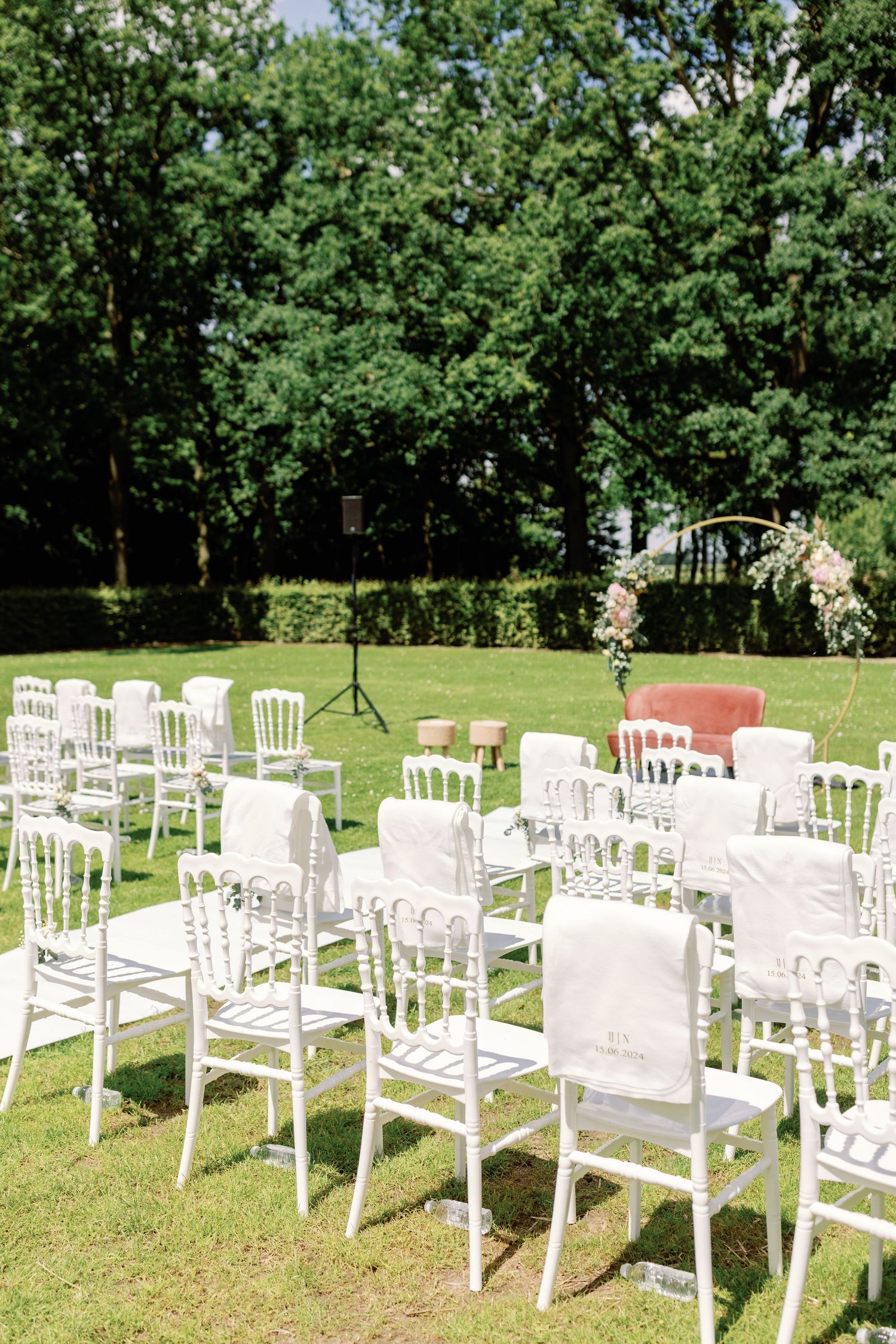 White wooden chairs arranged in rows on a green lawn for an outdoor wedding ceremony with trees in the background.