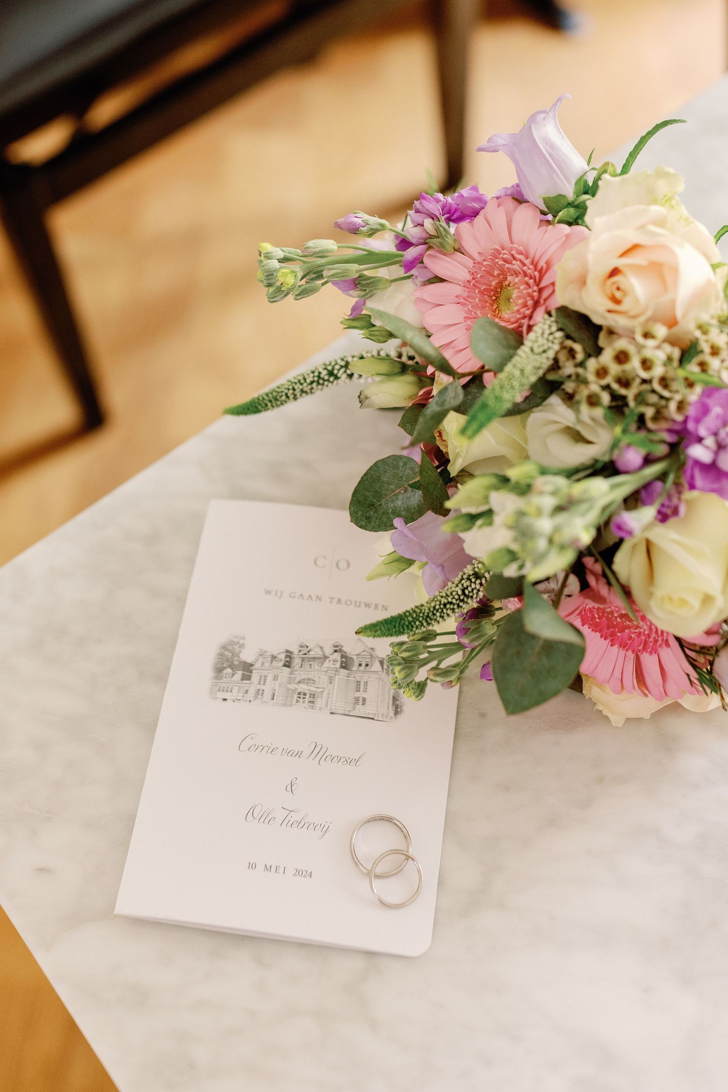 Wedding invitation with floral illustration and rings displayed alongside pink and peach flower arrangements on marble surface.