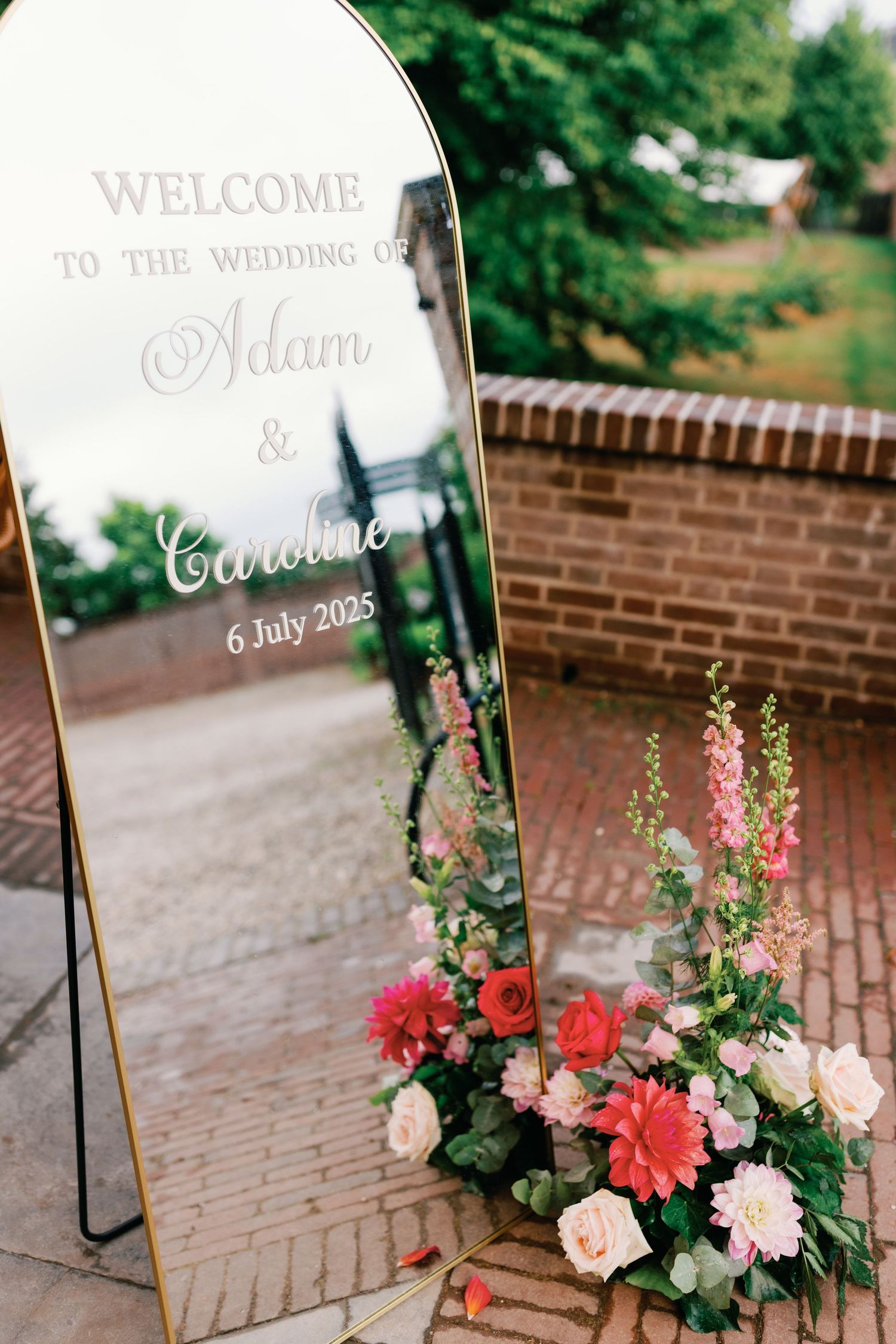 Elegant wedding welcome sign with floral arrangements featuring coral and pink blooms against a brick backdrop.