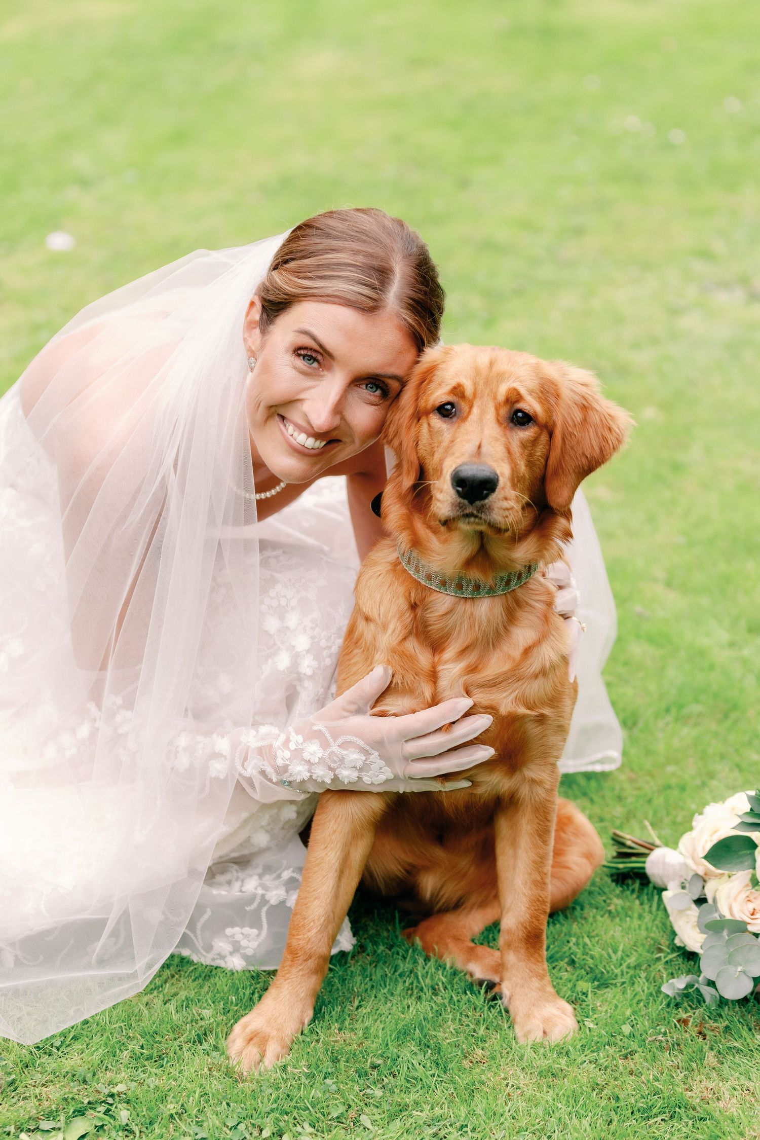 A golden retriever dog sits on grass next to someone in a white wedding dress holding a floral bouquet.