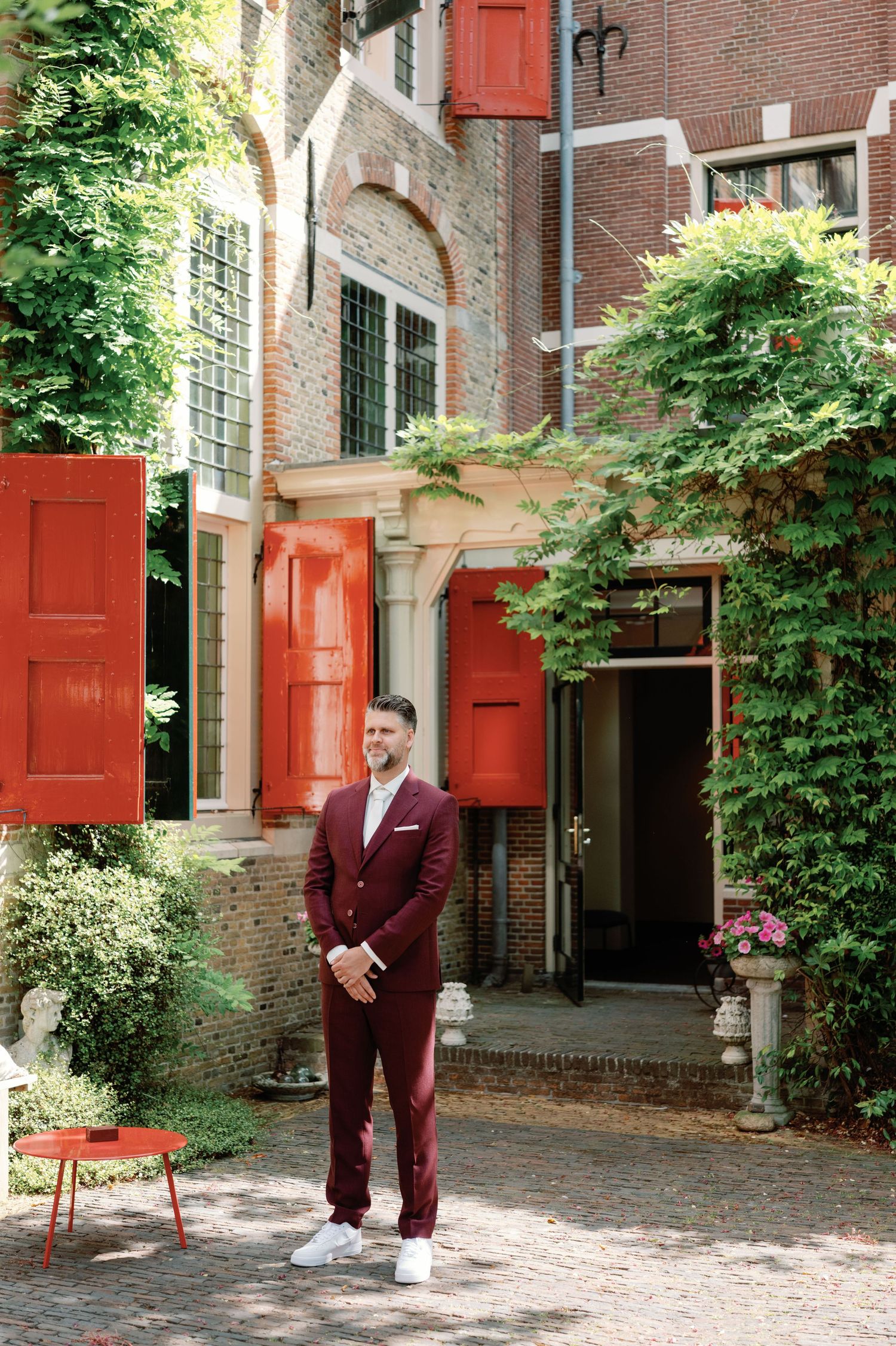 A burgundy-suited figure stands in a brick courtyard with red shutters and green foliage creating an elegant outdoor setting.