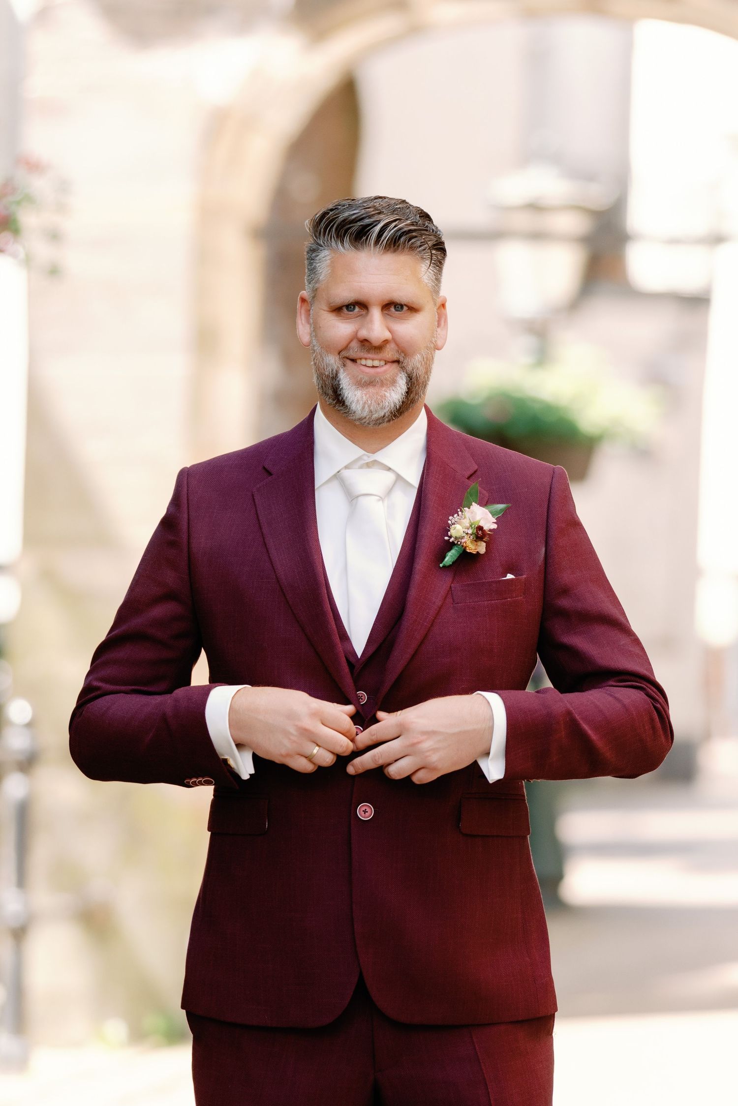 A groom in a burgundy suit with white tie and boutonniere buttons his jacket in an elegant outdoor setting.