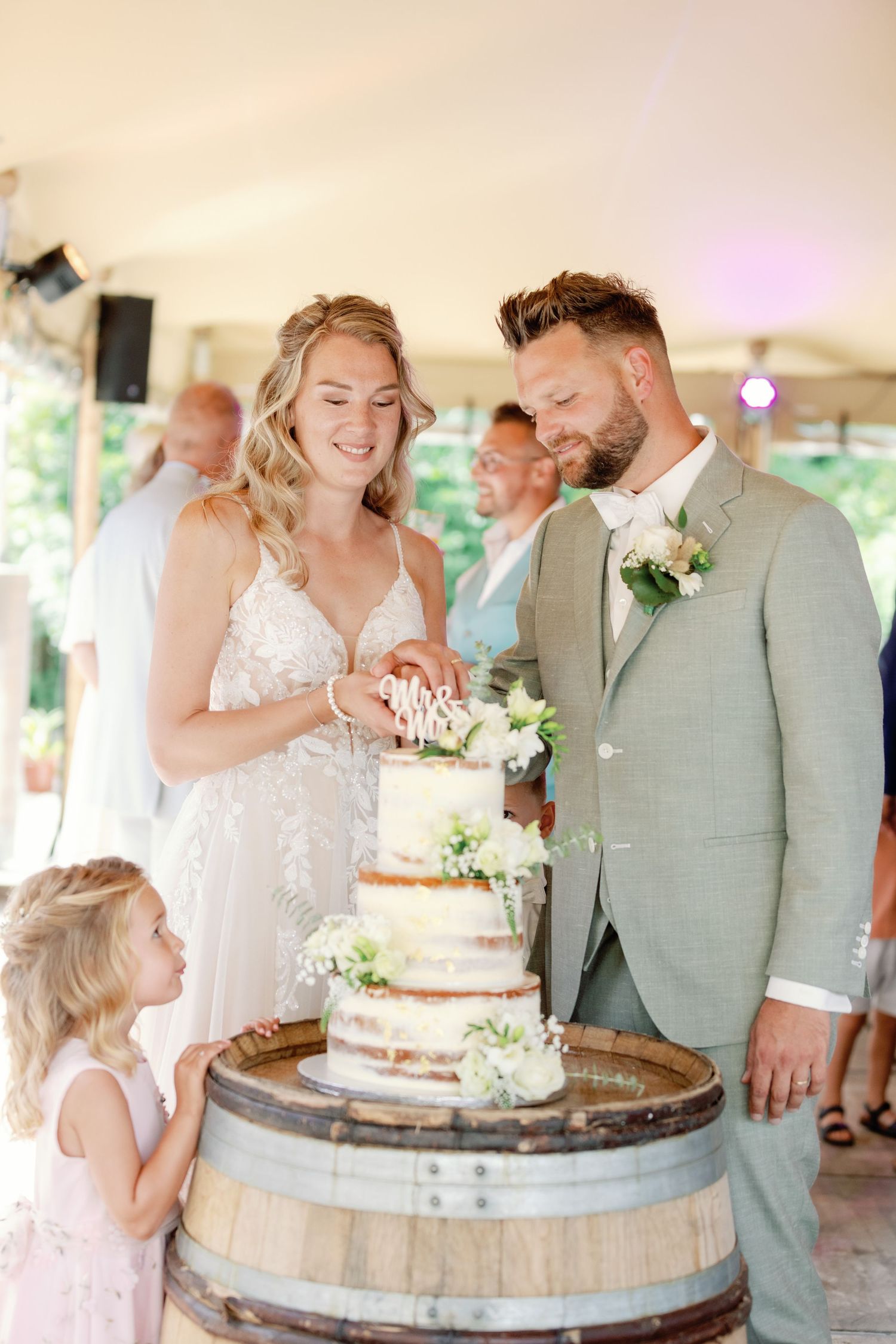 A couple shares a joyful moment cutting their rustic wedding cake displayed on a wooden barrel at an outdoor reception.