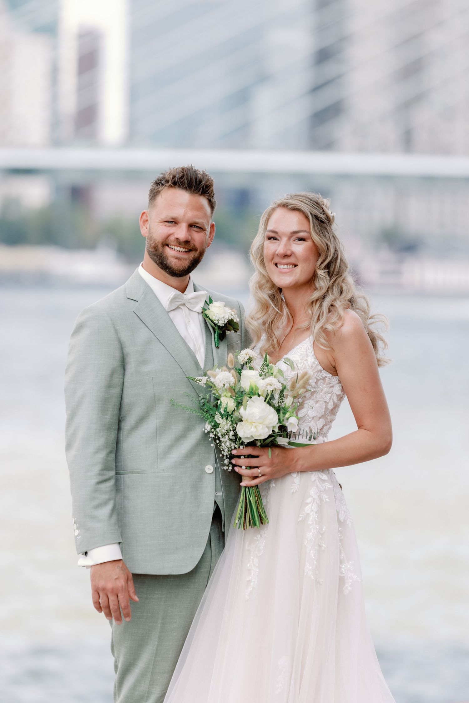 A couple in wedding attire share joyful moments together against a bright outdoor backdrop with white floral bouquet.