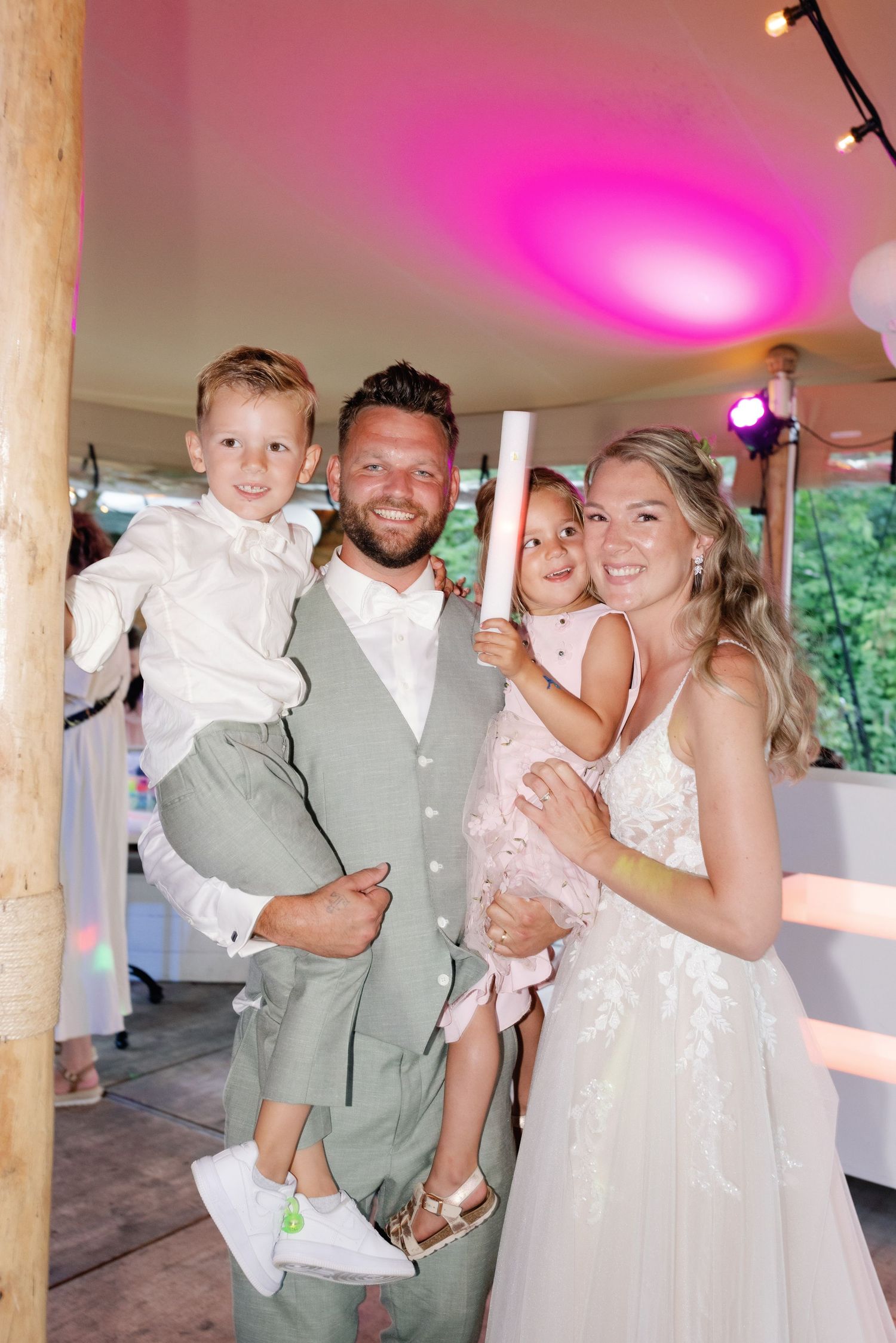Wedding couple dances with a young child during a reception under pink and purple lighting in a rustic venue.