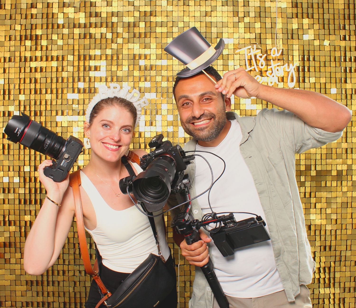 Two photographers smile holding cameras with props in front of a gold sequin backdrop.
