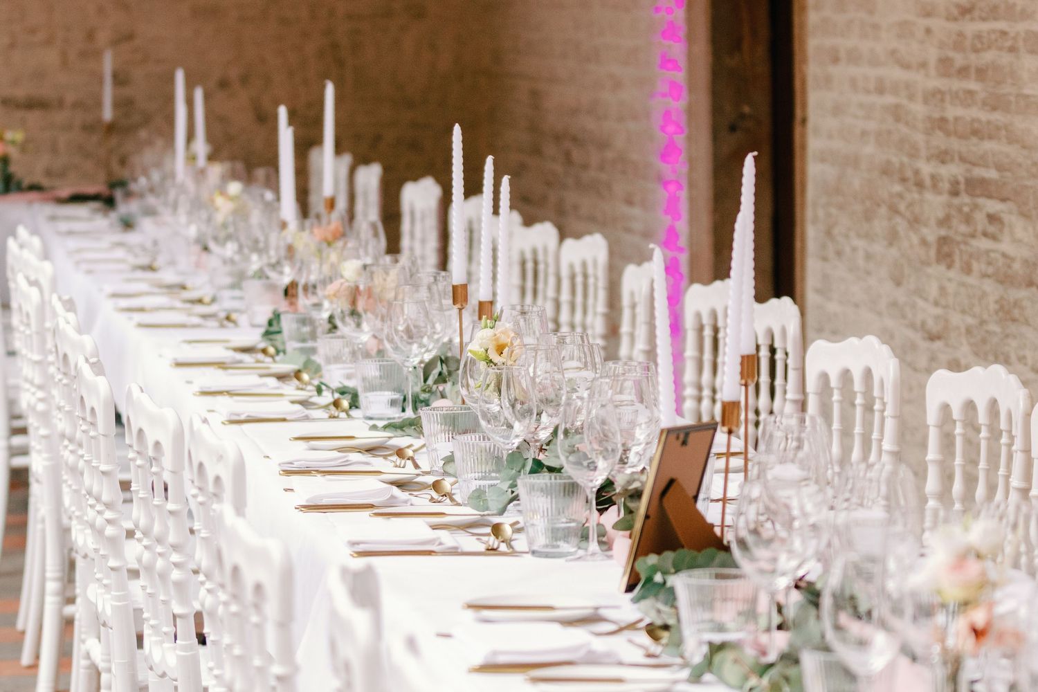 Long wedding reception table set with white chairs, glassware and floral centerpieces in an elegant brick-walled venue.