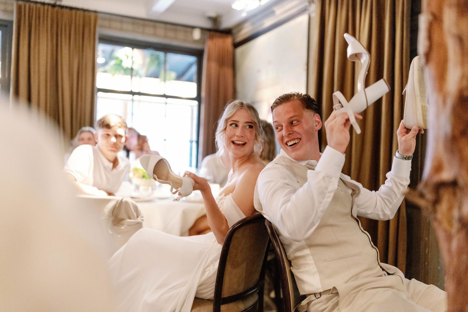 Wedding party celebrates joyfully with raised arms and excited expressions in a warmly lit indoor venue.
