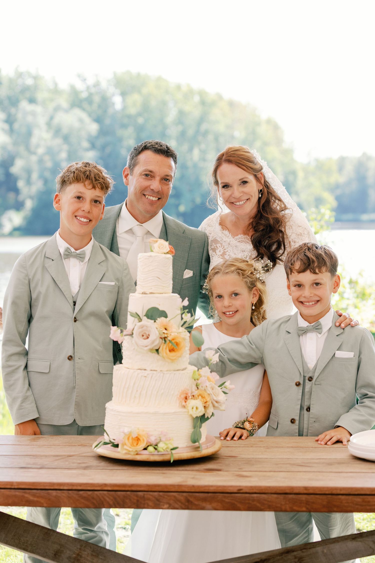 Wedding party in light gray suits gather around an elegant three-tiered white and peach cake at an outdoor reception.