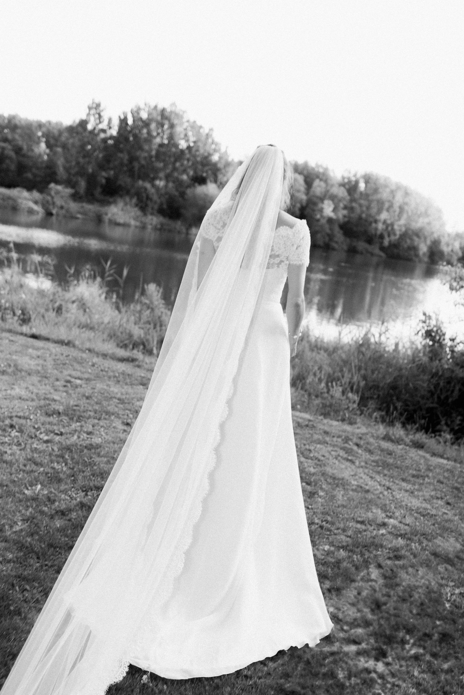 Black and white photo of a bride in a flowing white wedding gown with long veil standing by a lake.