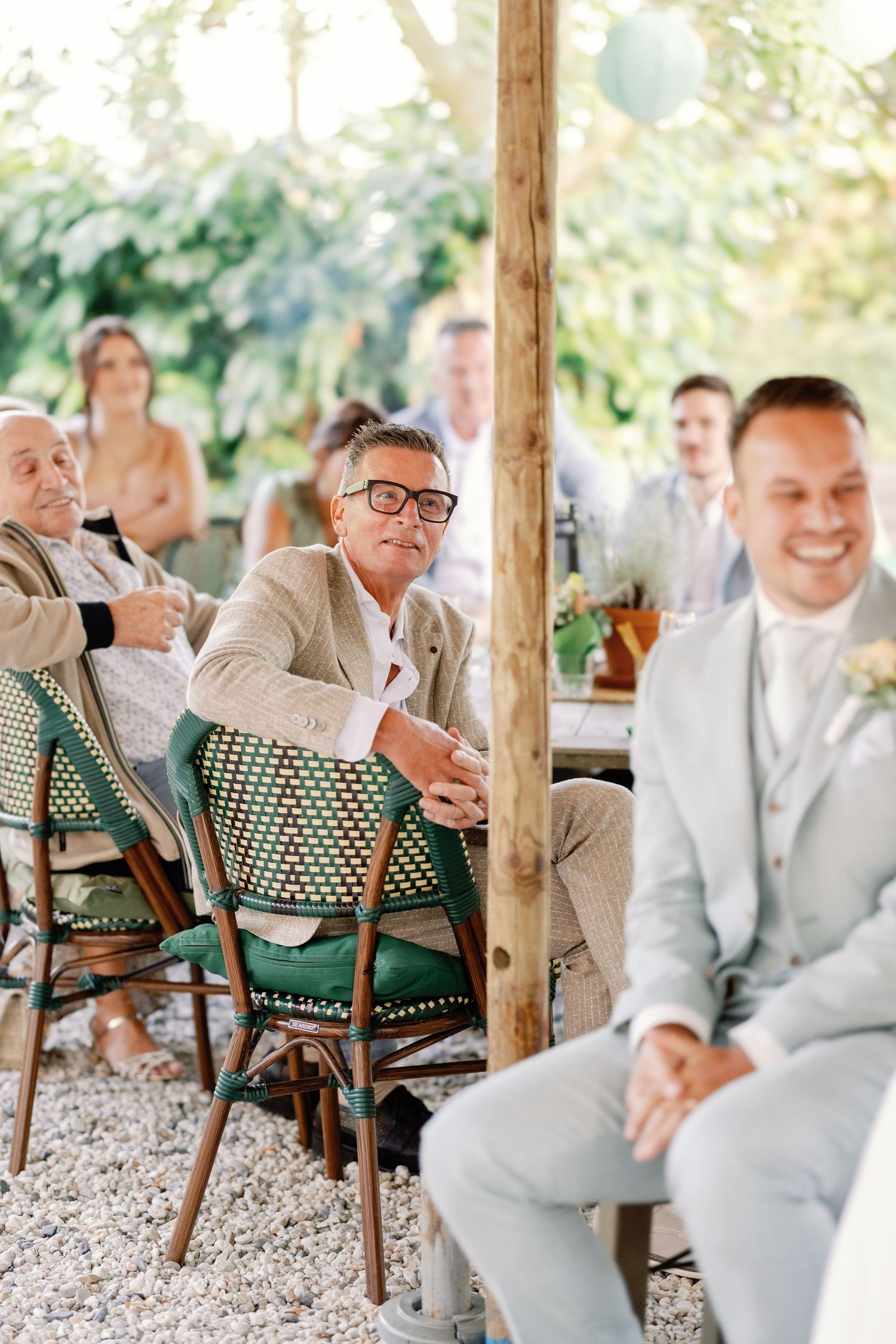 Wedding guests in light gray suits sit in bistro chairs under a rustic wooden pergola during an outdoor celebration.