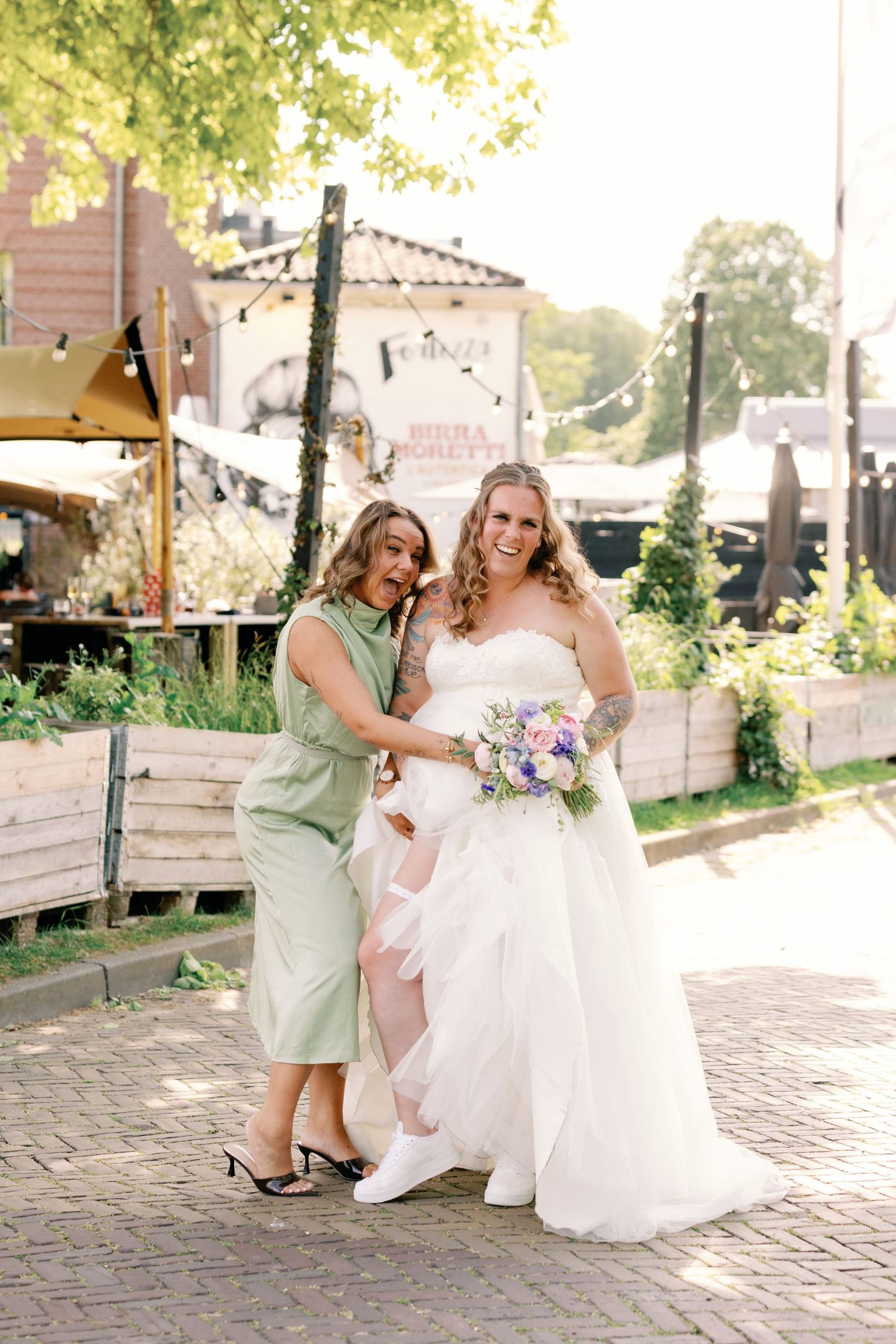 Bride in white gown holding bouquet poses with woman in green dress on brick path at outdoor venue.