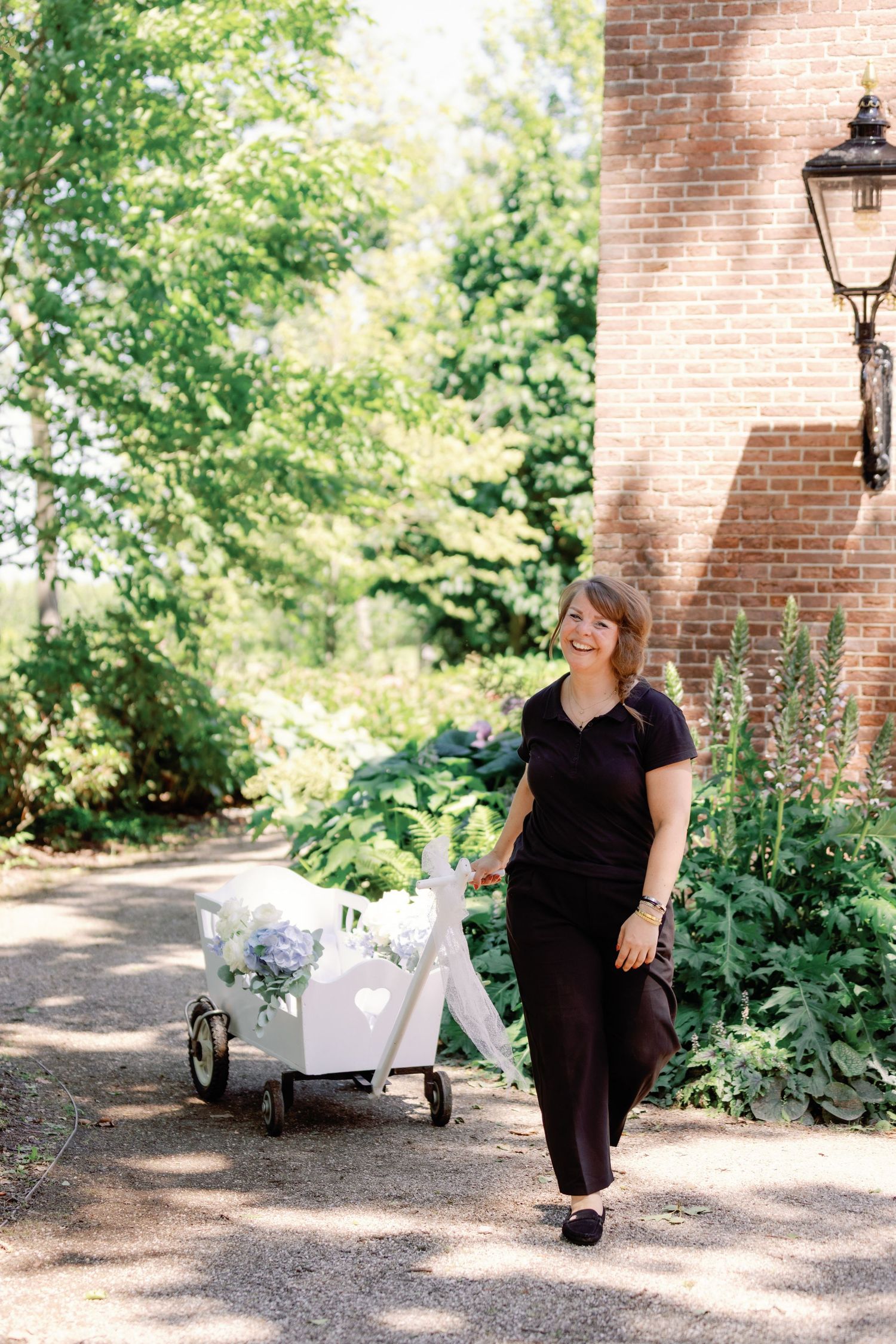 Woman in black outfit pulling a white floral cart along a garden path beside a brick building.